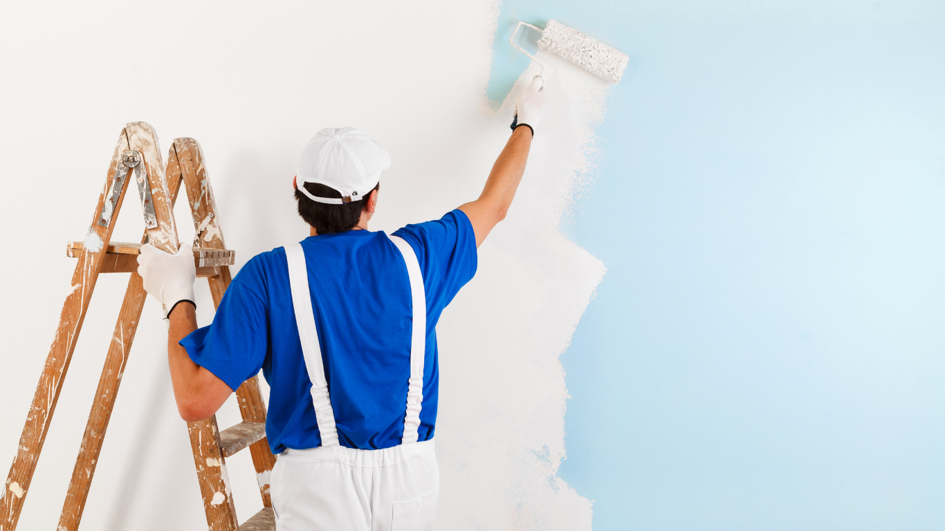 A painter in blue work clothes and a white cap paints a light blue wall white using a roller while standing on a ladder.