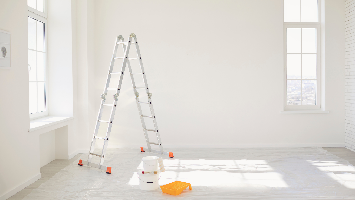 An aluminum stepladder stands in a bright, white empty room with a paint bucket and tray on a protective floor cover.
