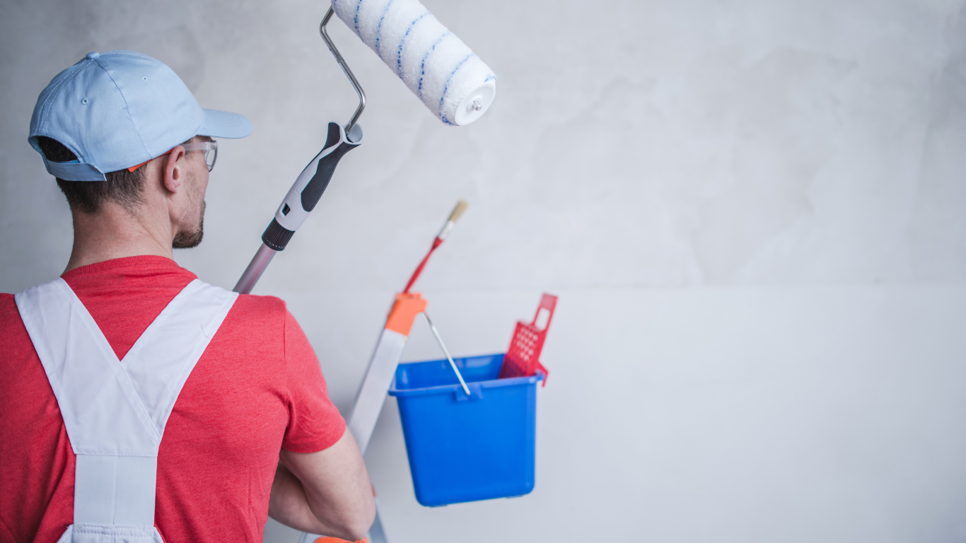 A painter in a red shirt and white overalls holds a paint roller in front of a blue bucket hanging on a white wall.
