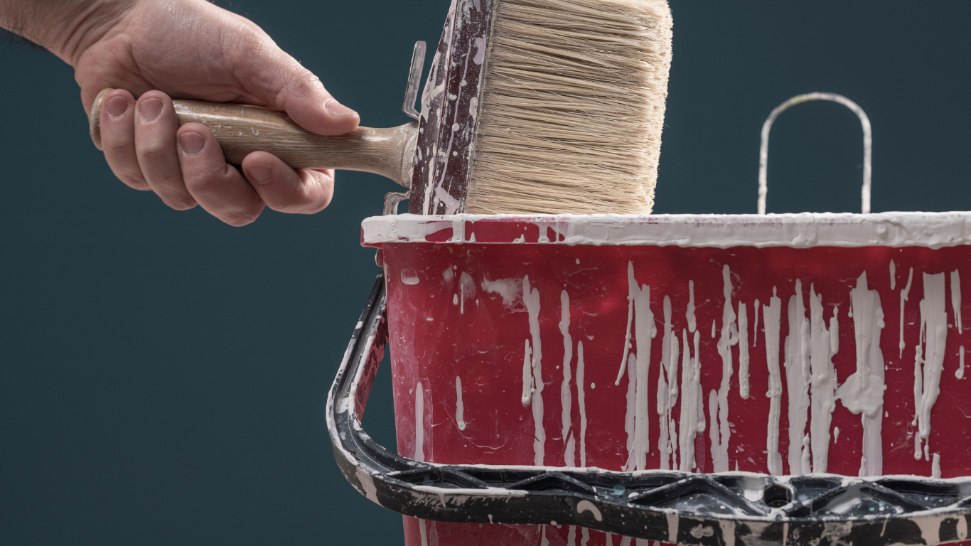 A hand holds a large block brush over a red plastic bucket dripping with white paint against a dark background.