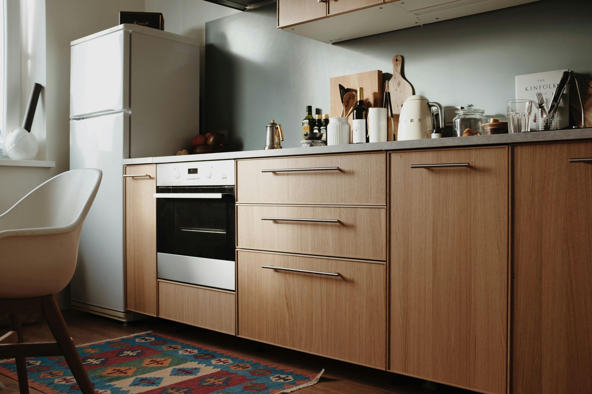 A kitchen with light wood cabinets, a white refrigerator, a built-in oven, and a patterned rug on the floor.