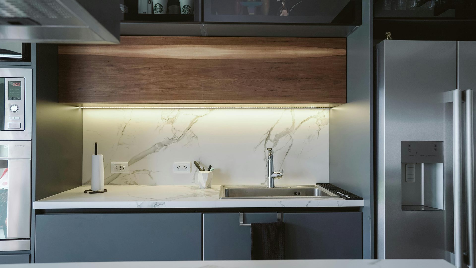 A modern kitchen with dark gray cabinets, wood accents, a white marble backsplash, and stainless steel appliances.