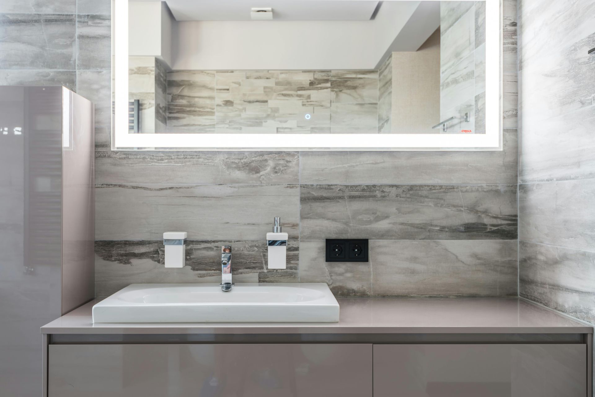 A modern bathroom vanity with a white rectangular sink, chrome faucet, illuminated mirror, and grey stone-look wall tiles.