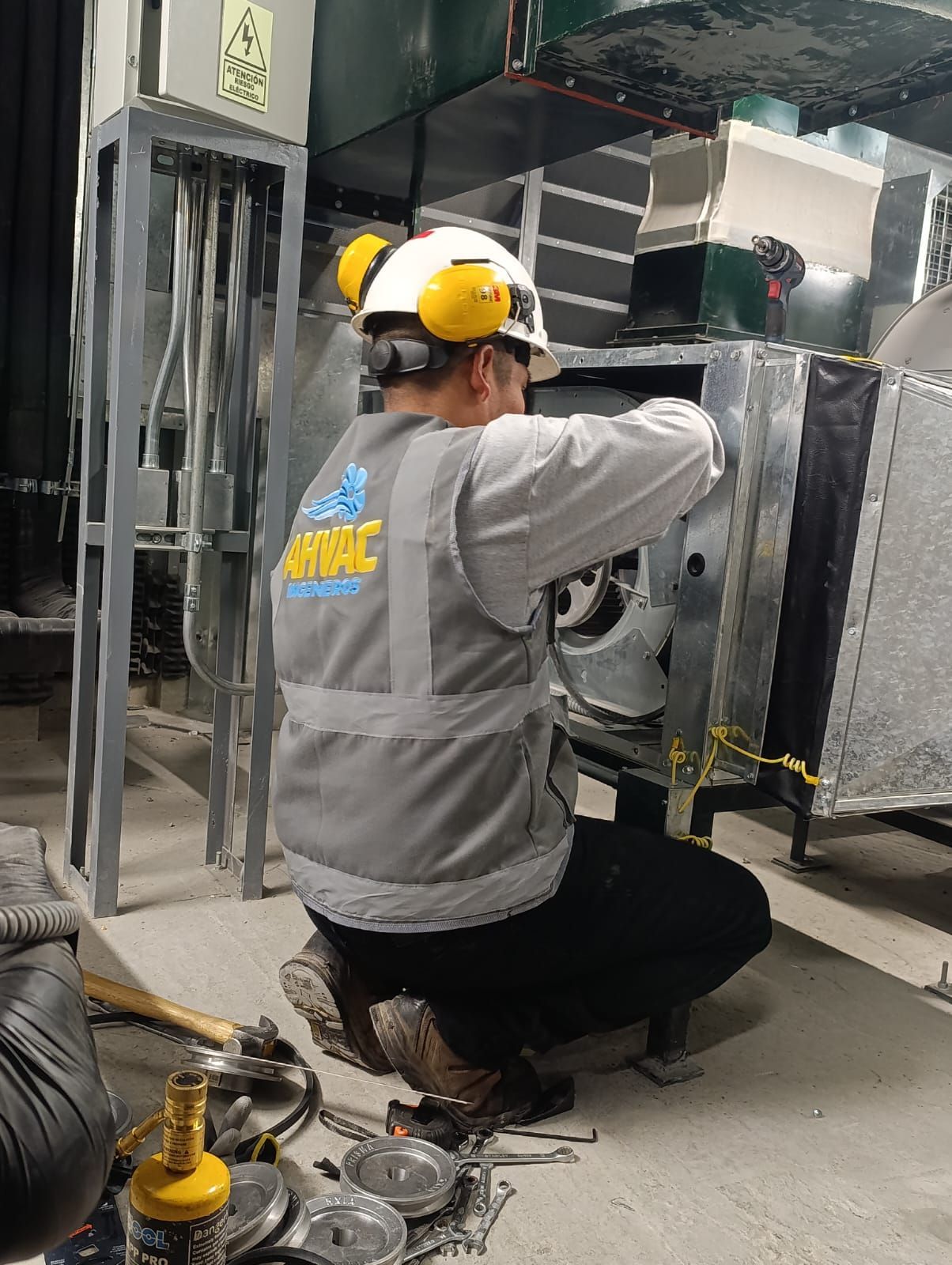Un trabajador con casco amarillo se arrodilla junto a un equipo industrial e inspecciona el cableado en un taller.