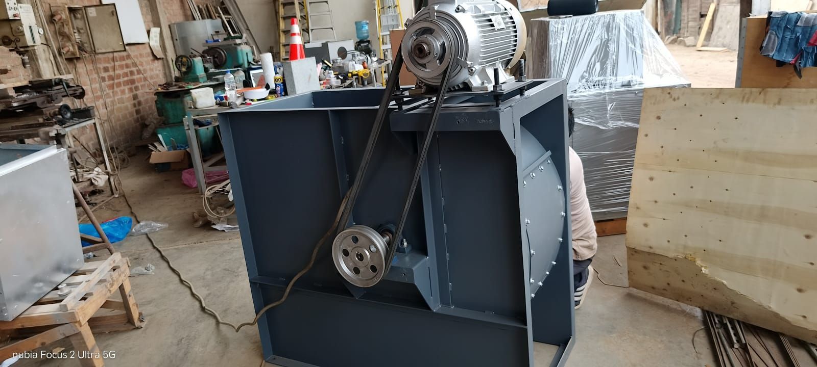 Máquina industrial con poleas y correas en un taller, con herramientas y materiales al fondo.