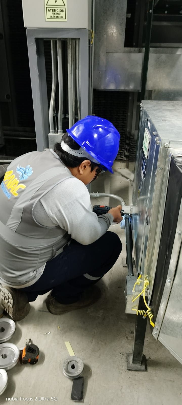 Un trabajador con casco azul se agacha para inspeccionar el cableado de un panel eléctrico industrial.