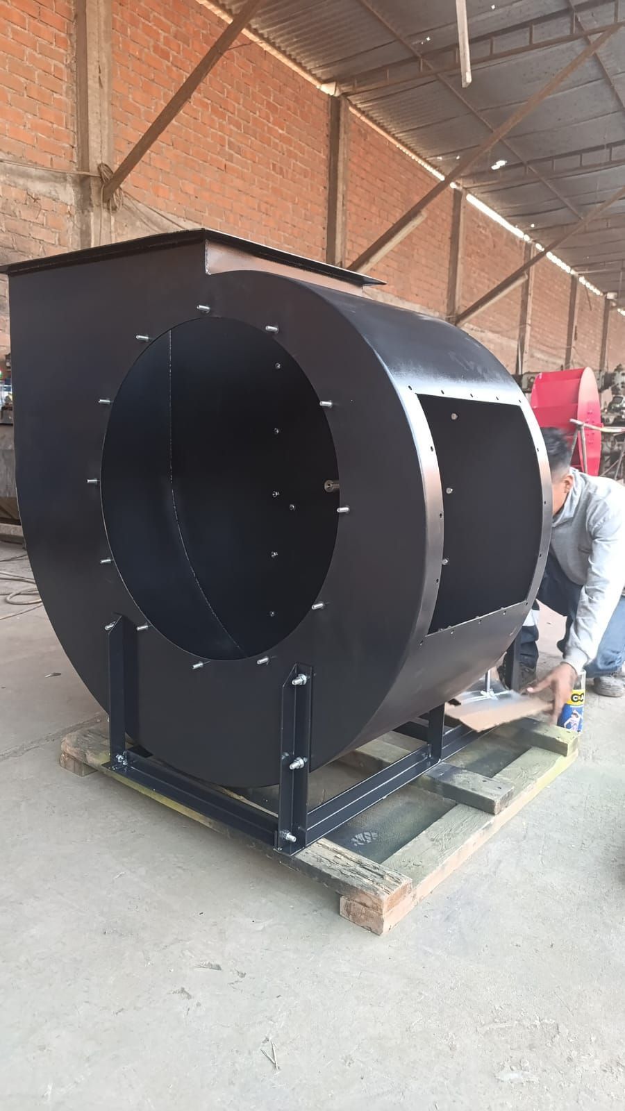 Carcasa de ventilador industrial grande de color negro sobre un palé en un almacén, con un trabajador ensamblándola.
