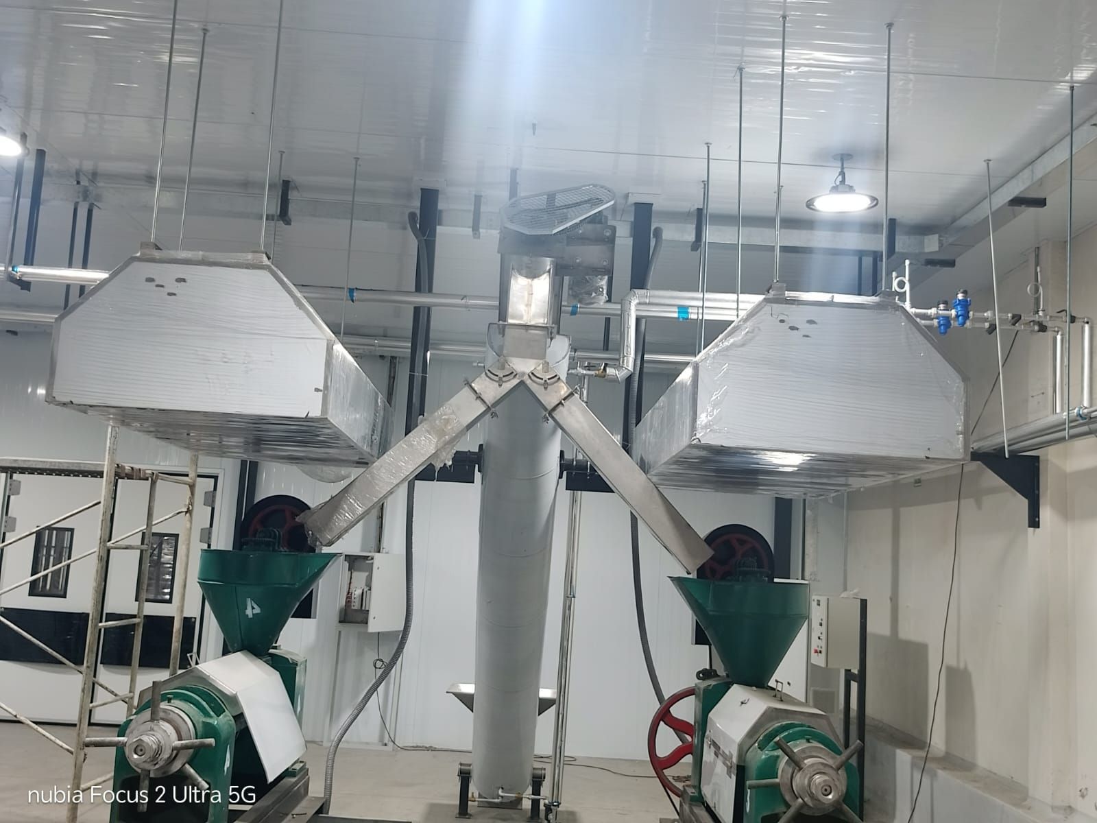 Sala industrial con dos trabajadores manejando maquinaria verde bajo conductos y rejillas de ventilación metálicas.