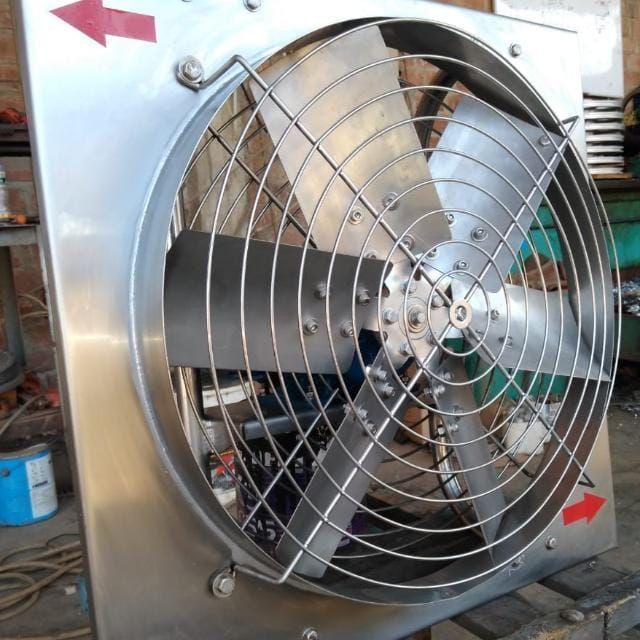 Ventilador industrial de pared con rejilla metálica y flechas rojas indicadoras de dirección sobre un panel plateado.