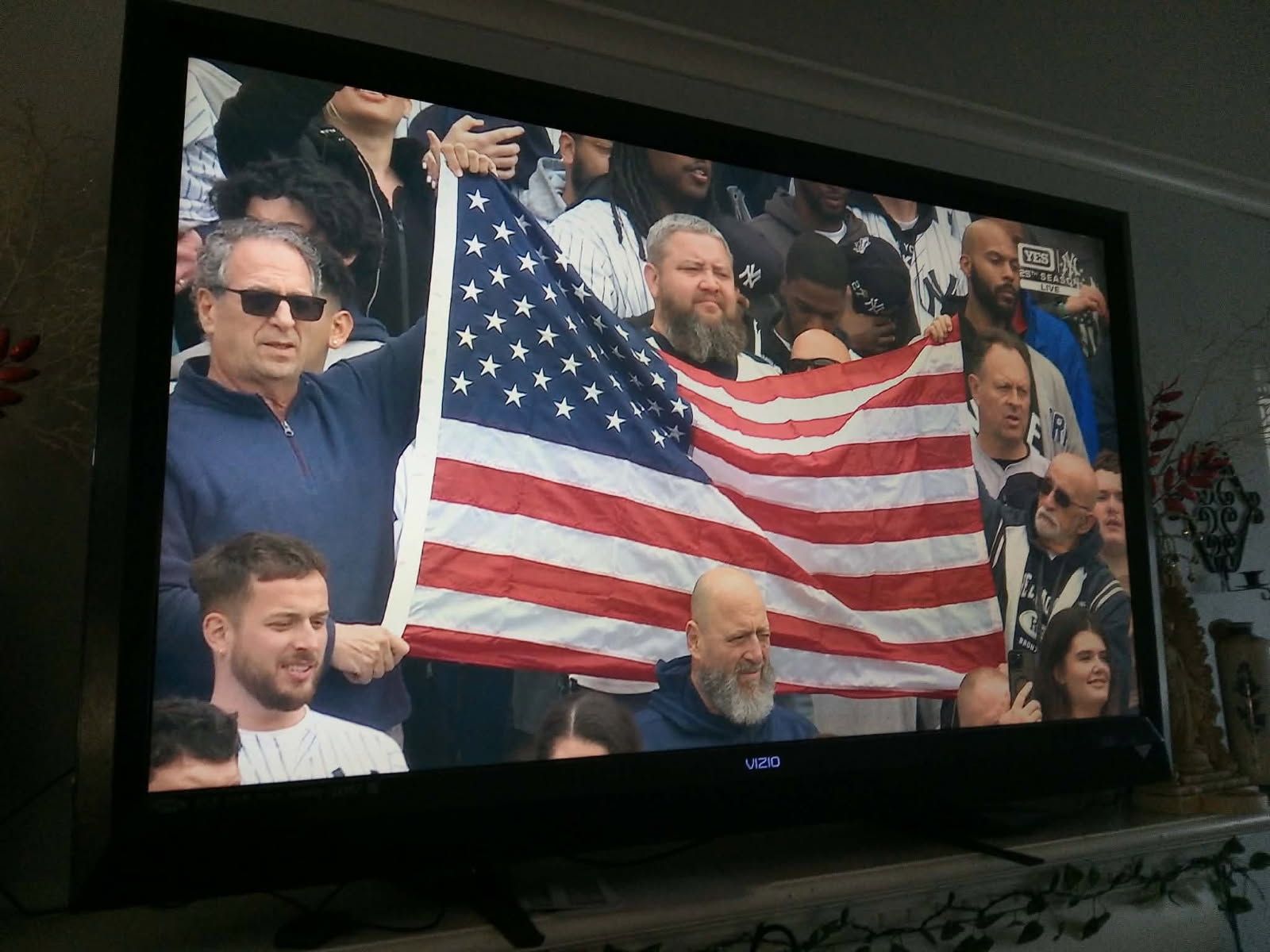 Crowd at a stadium holding a large American flag, with spectators seated behind them.