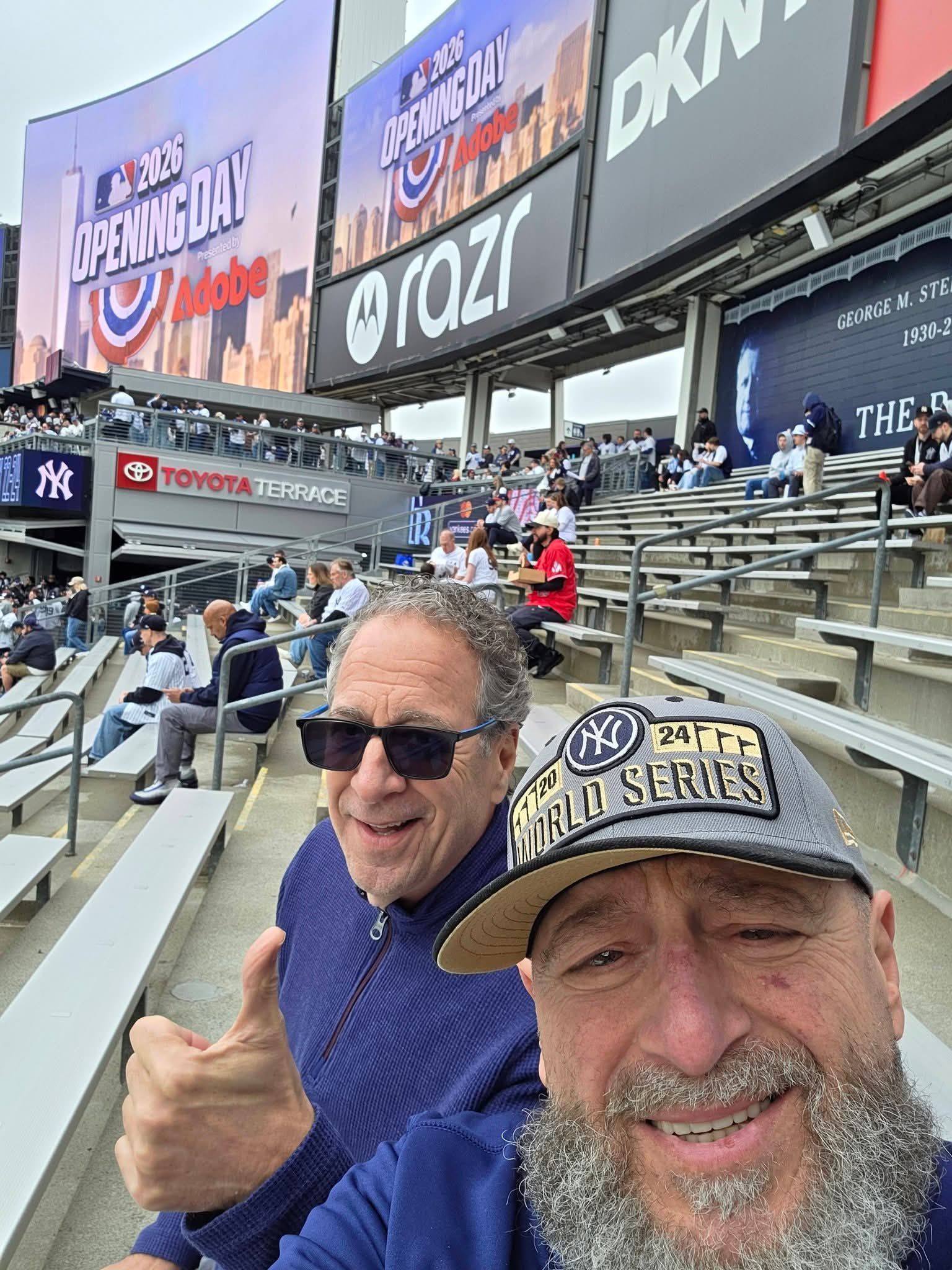 Two smiling fans in baseball caps at a stadium, with one giving a thumbs-up in the stands.