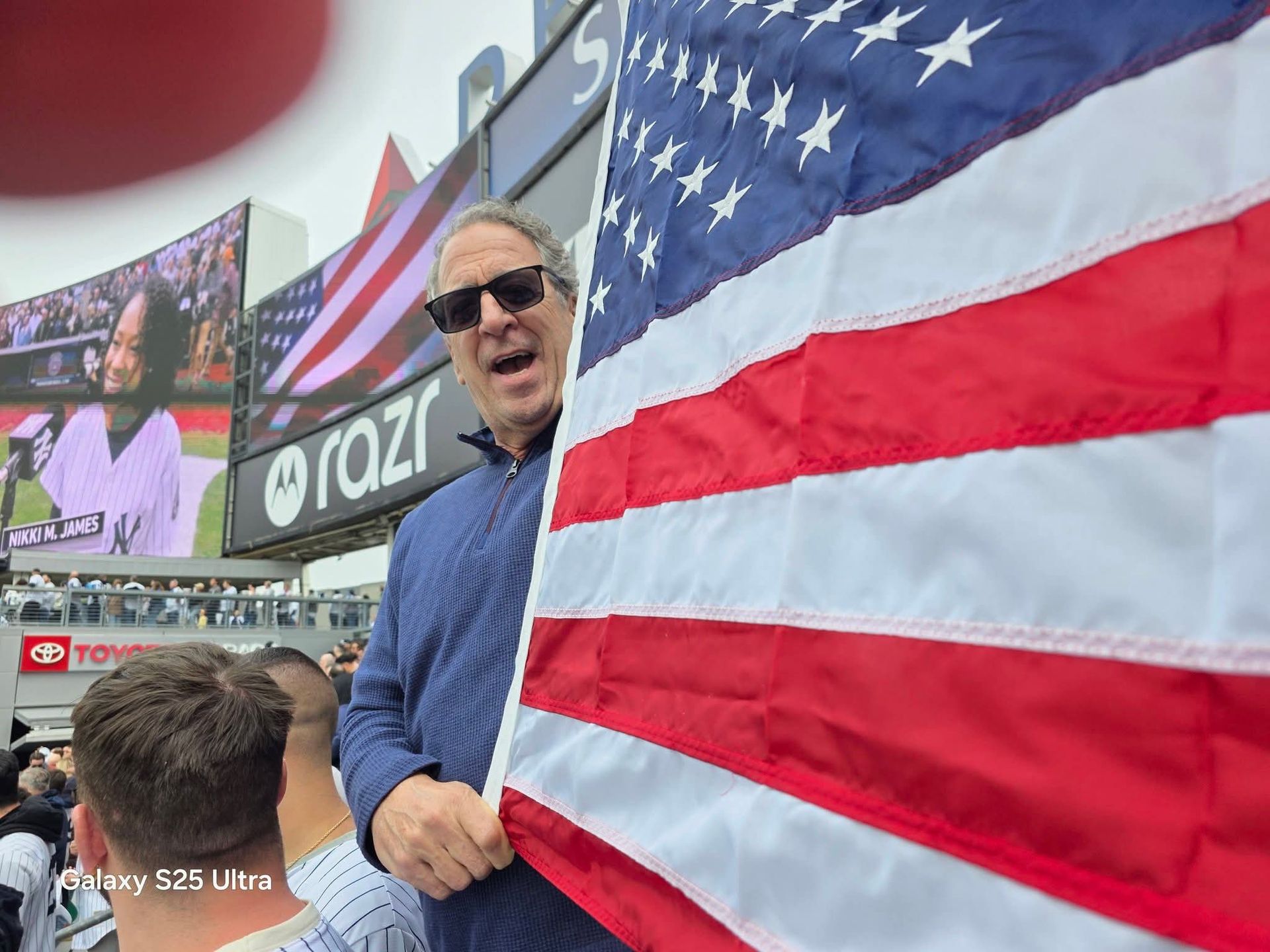 Man waving a large American flag at a crowded outdoor event with billboards in the background