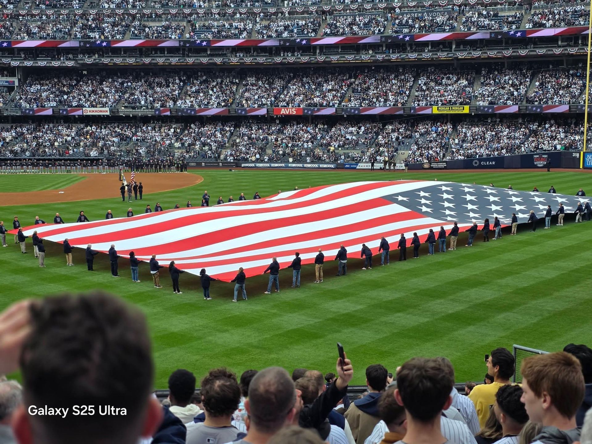Large American flag held on a baseball field during a stadium ceremony, with a crowd in the stands