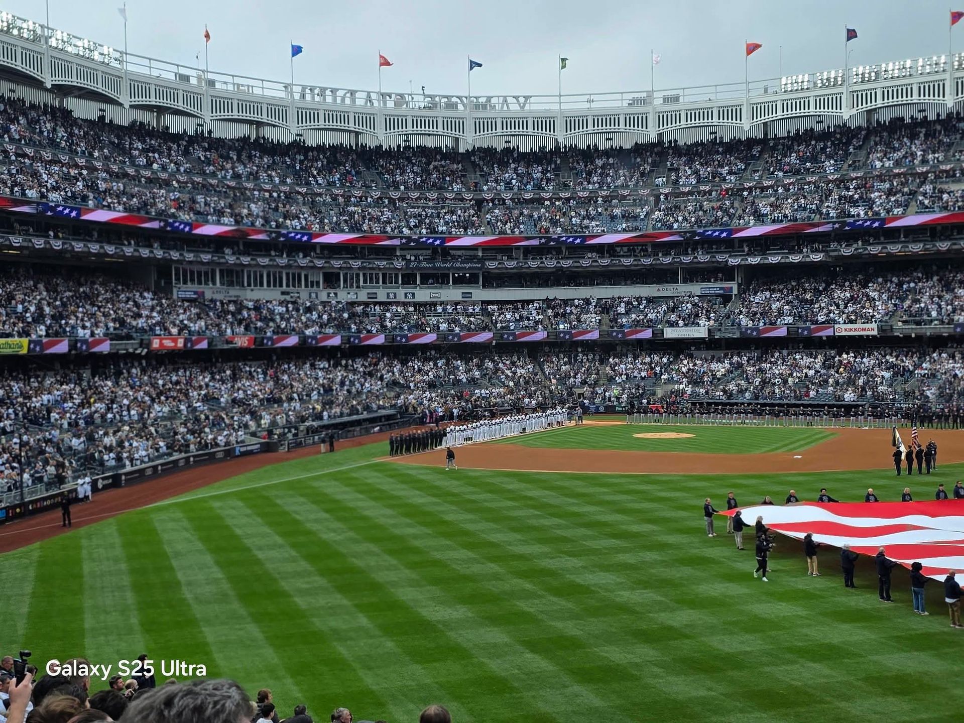 Crowded baseball stadium during a pregame ceremony, with a large U.S. flag on the field