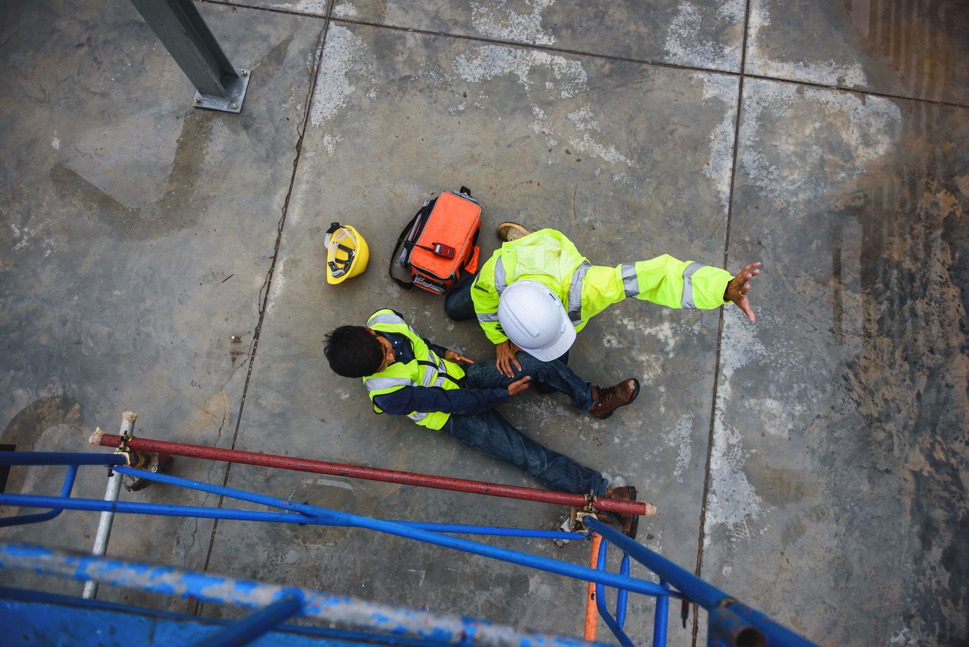 Two workers in neon safety gear kneel on a wet concrete surface beside a blue railing and orange equipment pack.