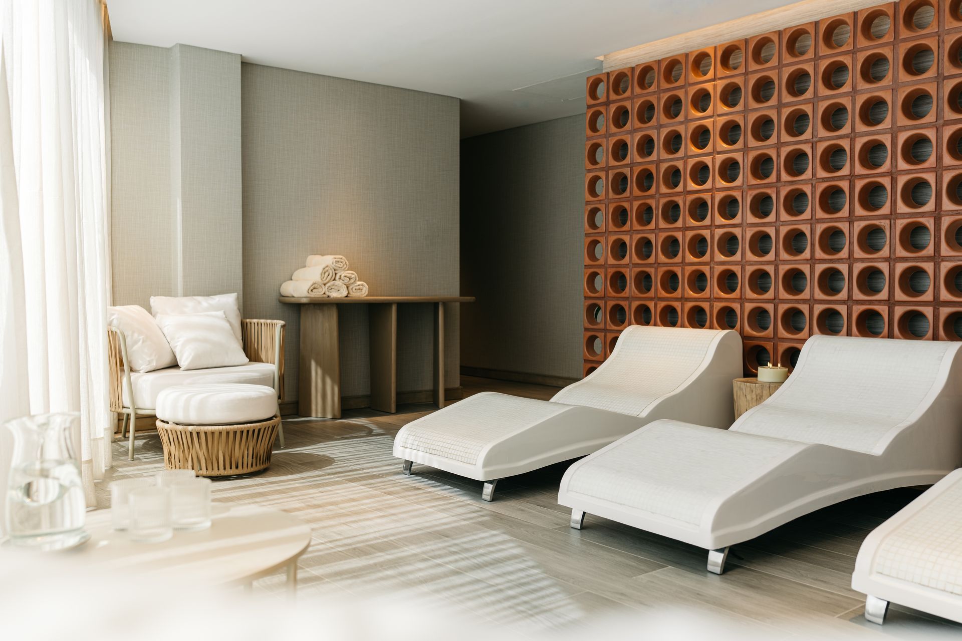 Sala de relajación con tumbonas blancas, pared de terracota texturizada y cortinas transparentes.