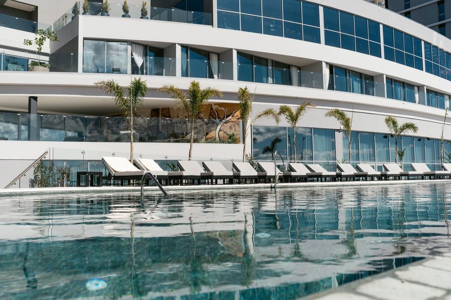 Piscina al aire libre con borde curvo, camastros blancos, palmeras y barandilla de cristal.