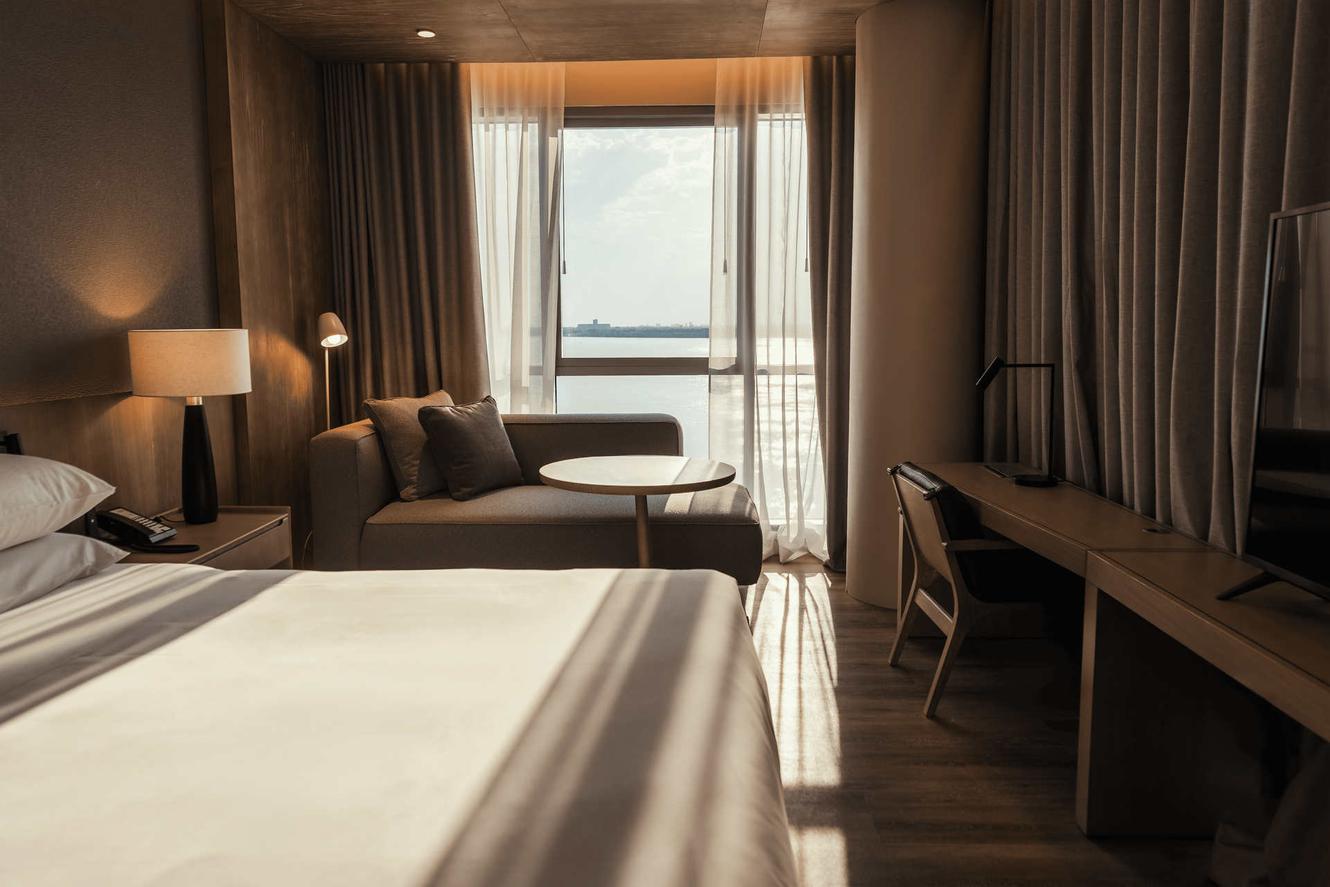 Habitación de hotel con cama, sofá, escritorio, ventana con cortinas transparentes e iluminación cálida.