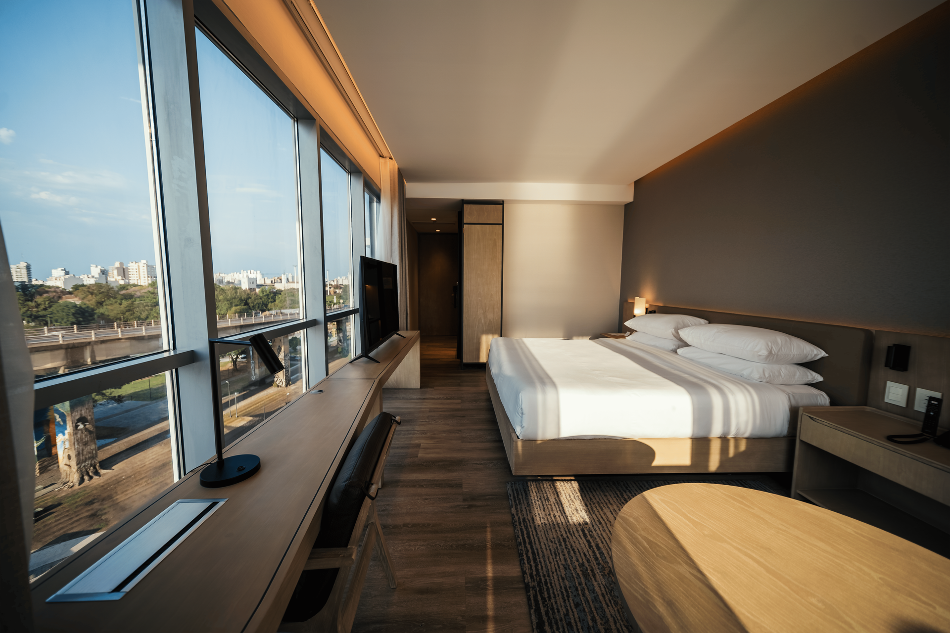 Habitación de hotel moderna con grandes ventanales con vistas a la ciudad, cama tamaño king, escritorio y combinación de colores neutros.