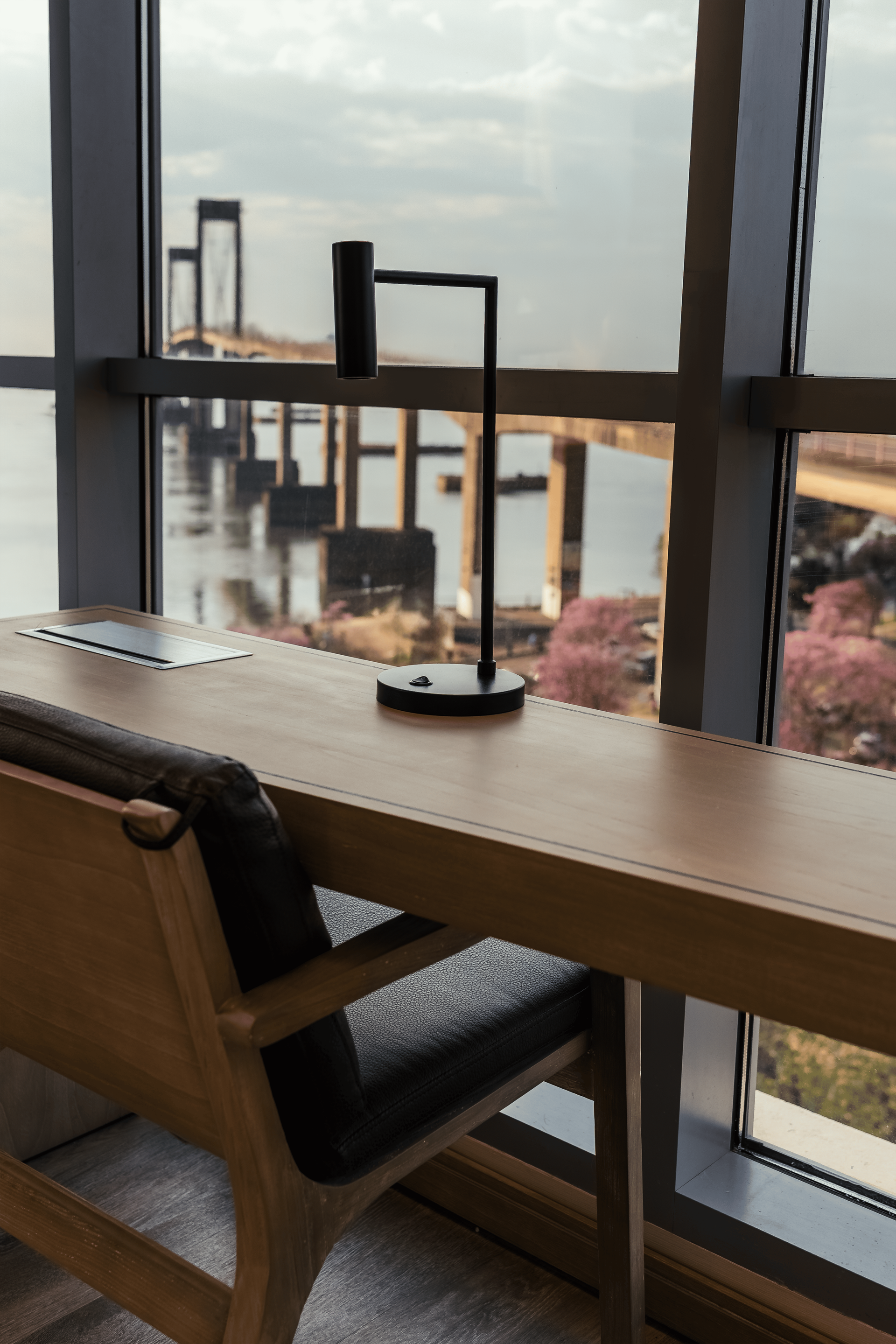 Escritorio junto a una ventana con vista al puente. Lámpara negra, silla de madera, escritorio con papeles y árboles con flores rosas.