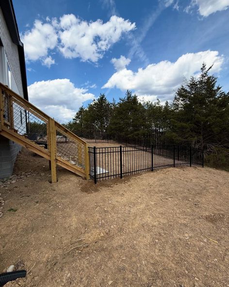 Gravel yard beside a house with a wooden staircase, black metal fence, trees, and blue sky with clouds