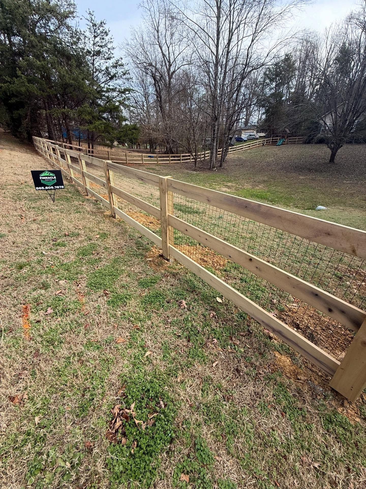 Wooden split-rail fence beside a grassy field with trees and a small yard sign.