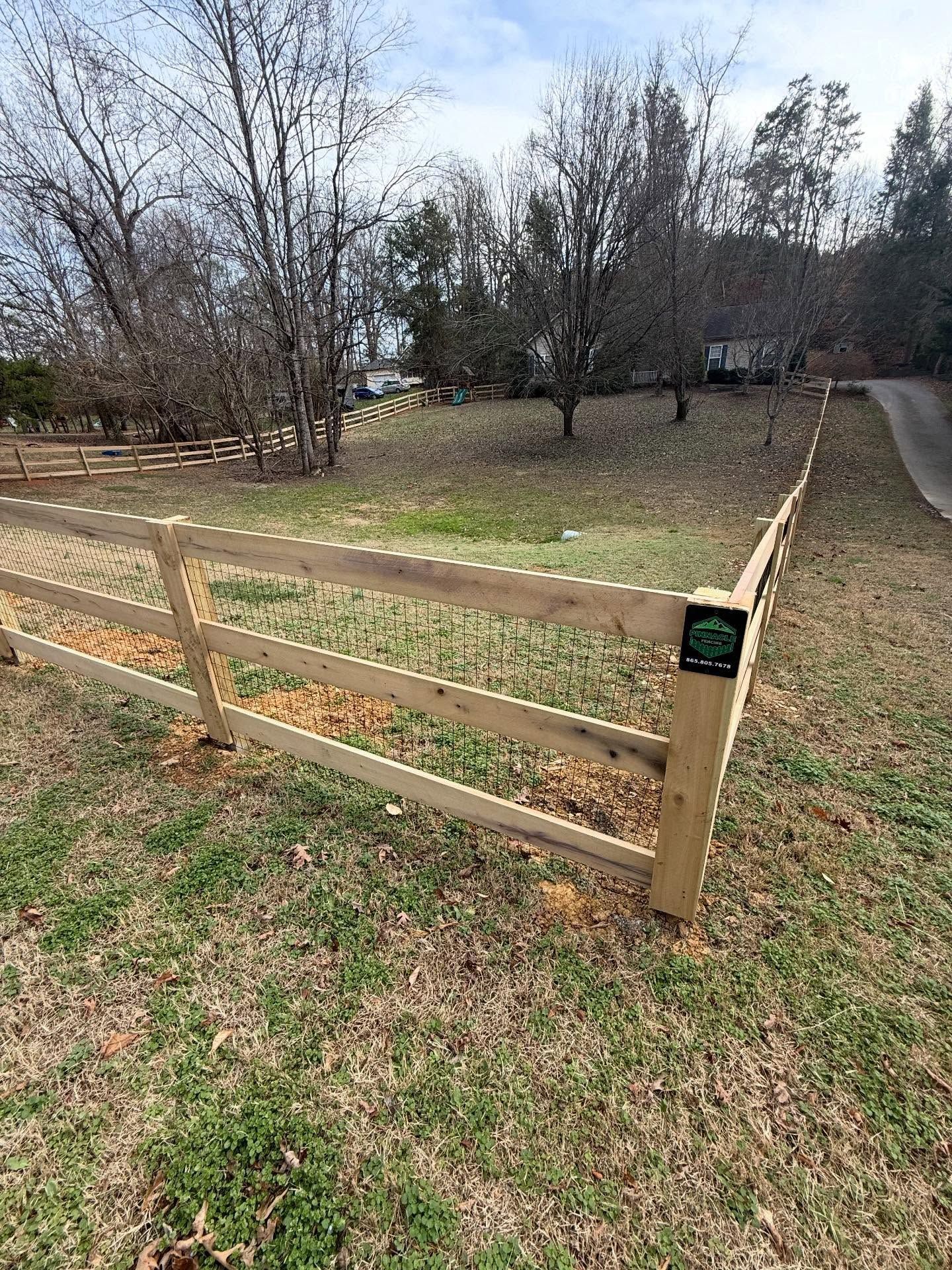 Wooden fence corner bordering a grassy yard with leafless trees and a paved path beside it