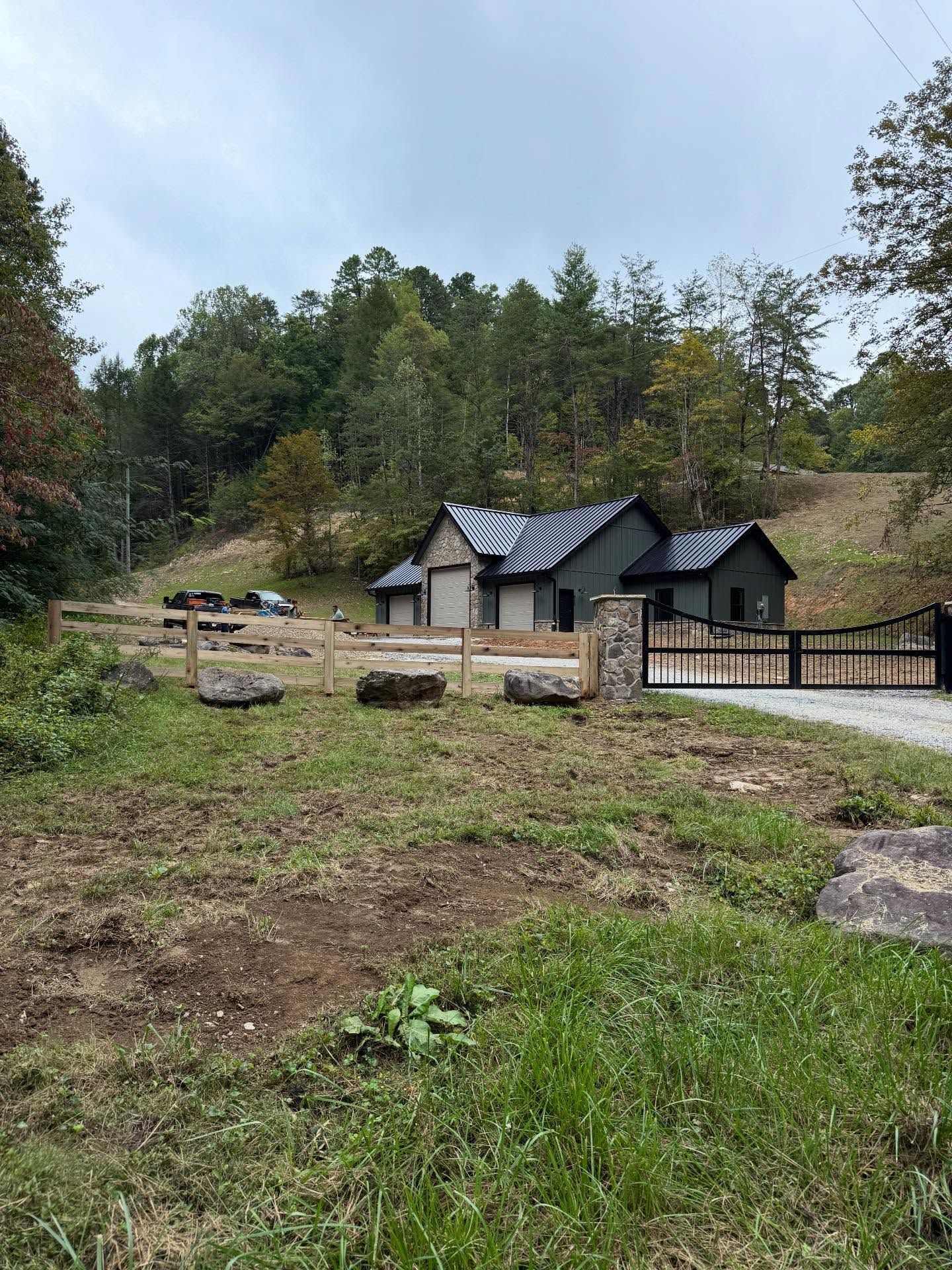Rural cabin with black roof and stone chimney behind a fenced gravel driveway and grassy foreground.