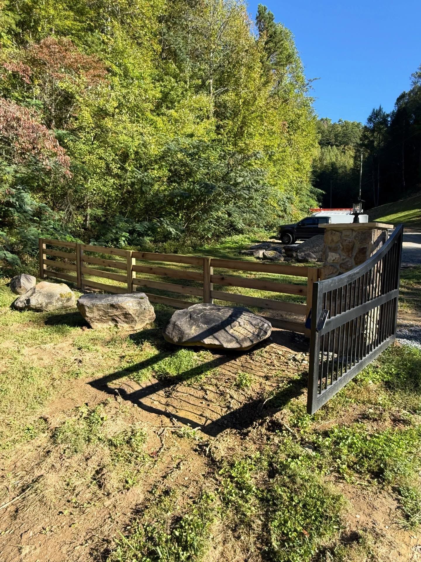 Wooden fence and gate beside large rocks on a grassy path in a sunny wooded area