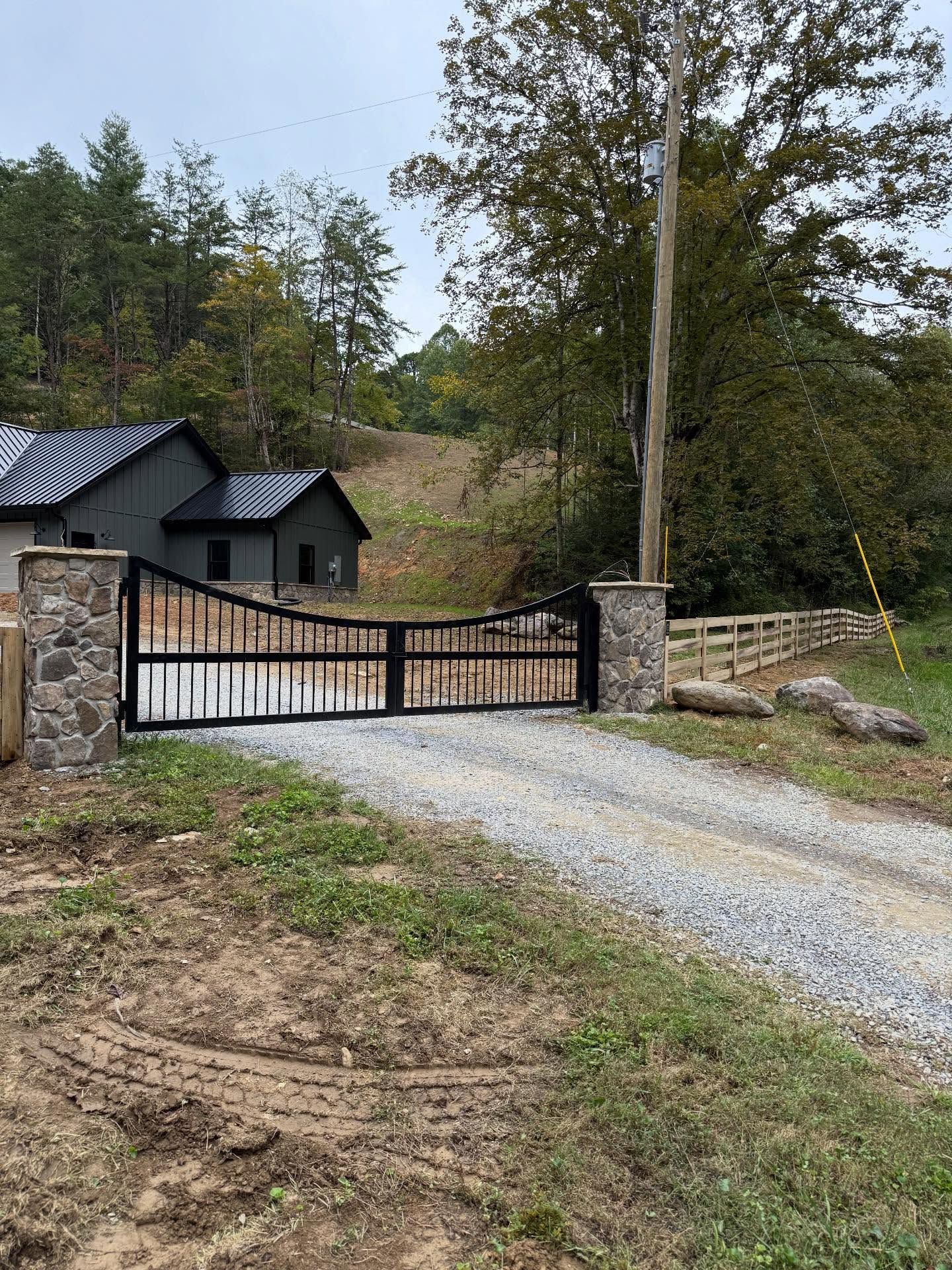 Gravel driveway with black gate and stone pillars leading to a dark gray house in a wooded rural setting