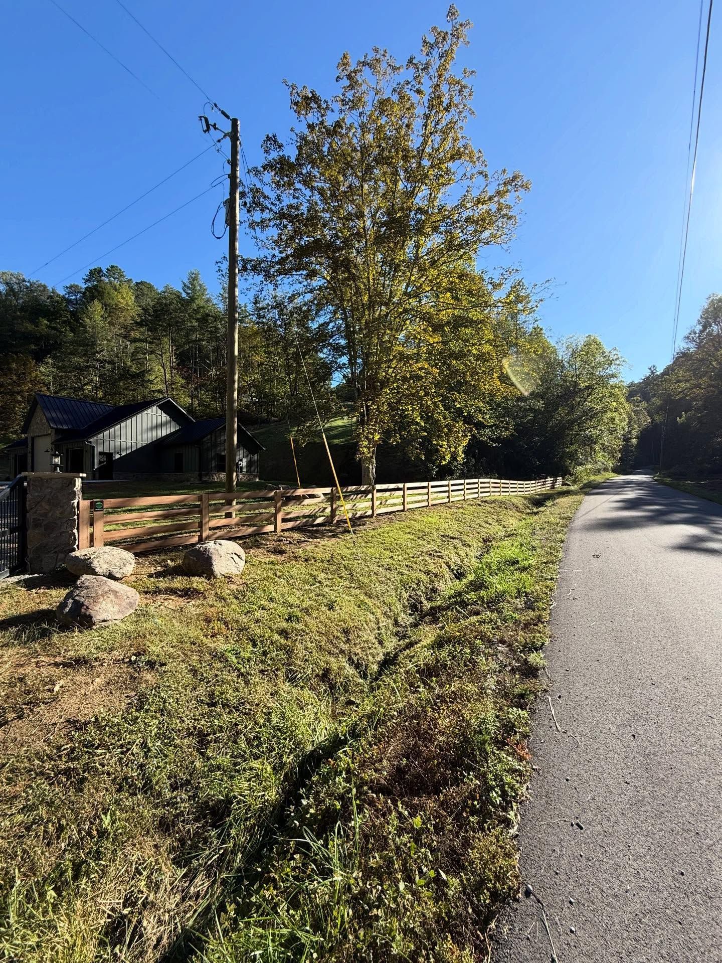 Rural roadside with a tree, wooden fence, and stone wall under a clear blue sky