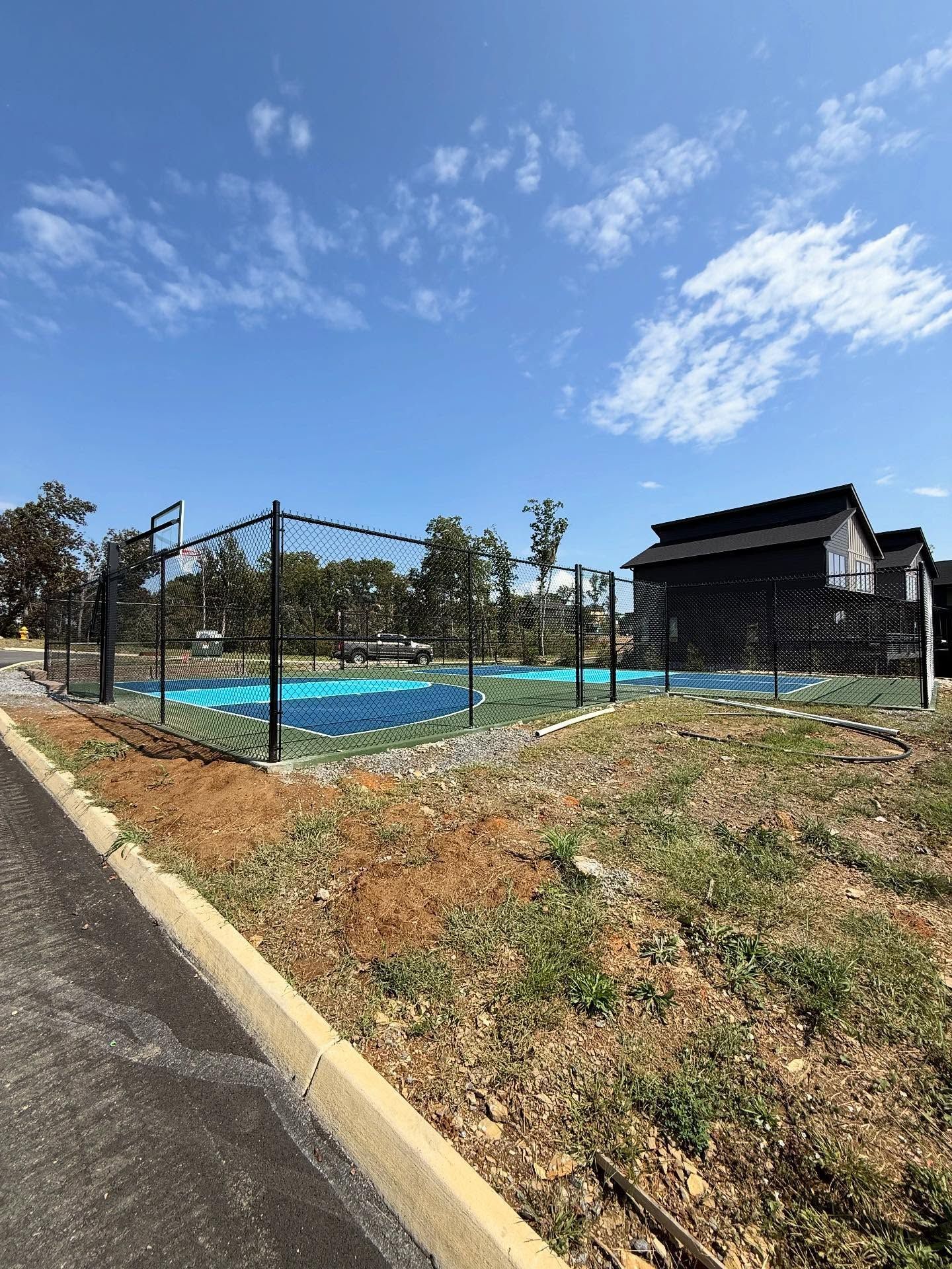 Outdoor tennis court with black fence, blue surface, and a small clubhouse under a partly cloudy sky