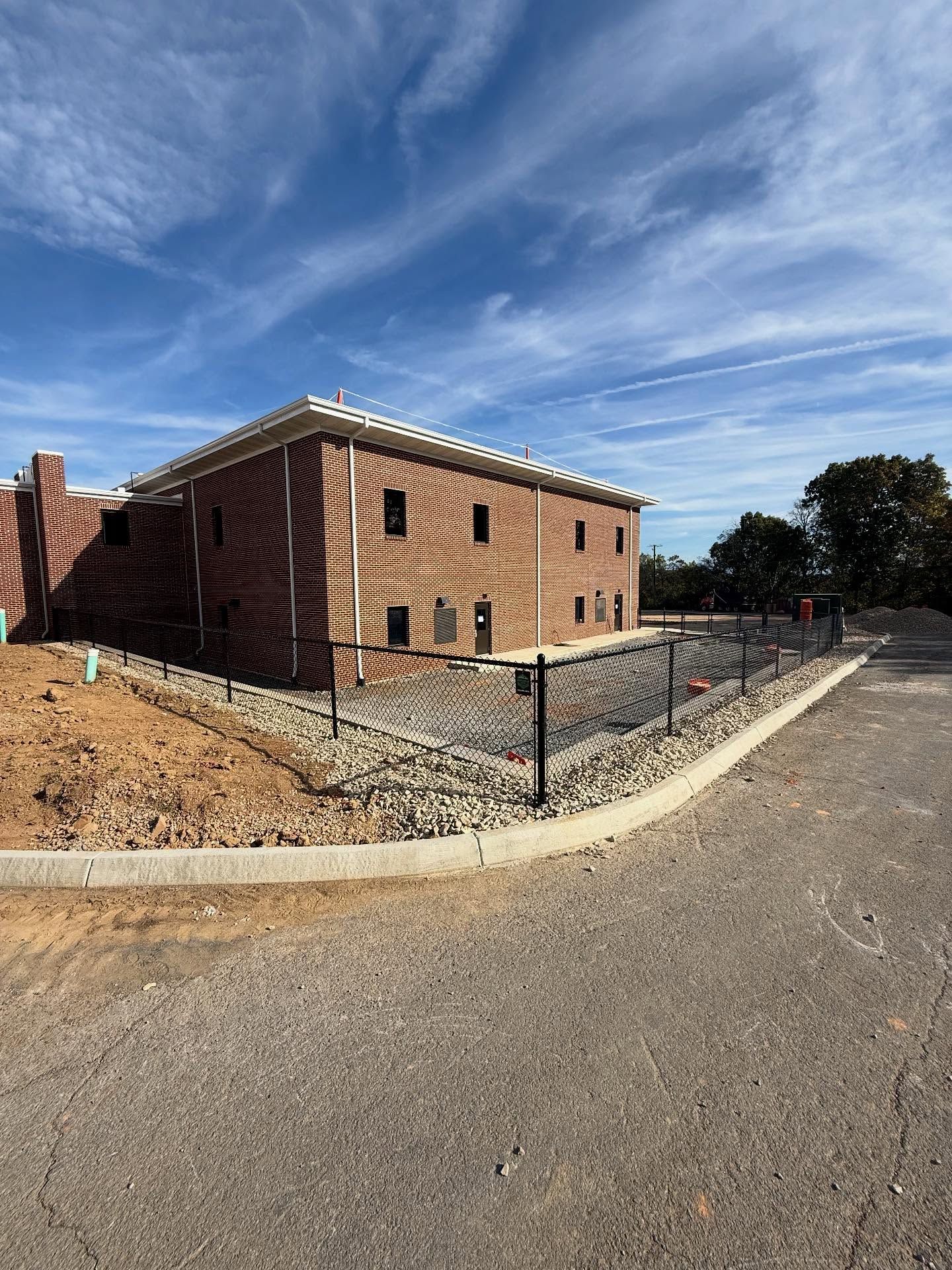 Brick building under construction behind a chain-link fence on a gravel lot, with a blue sky overhead