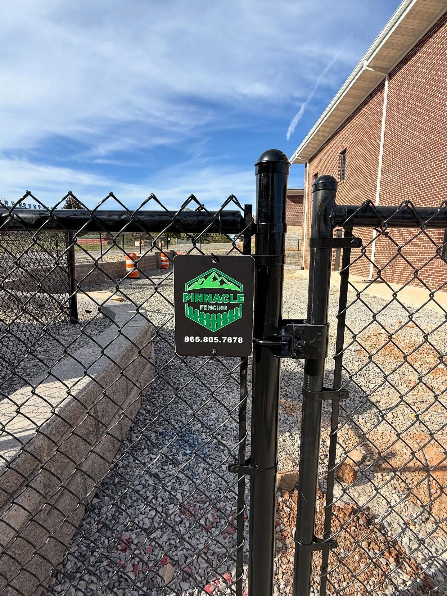 Chain-link fence gate with a green security sign beside a brick building under a blue sky
