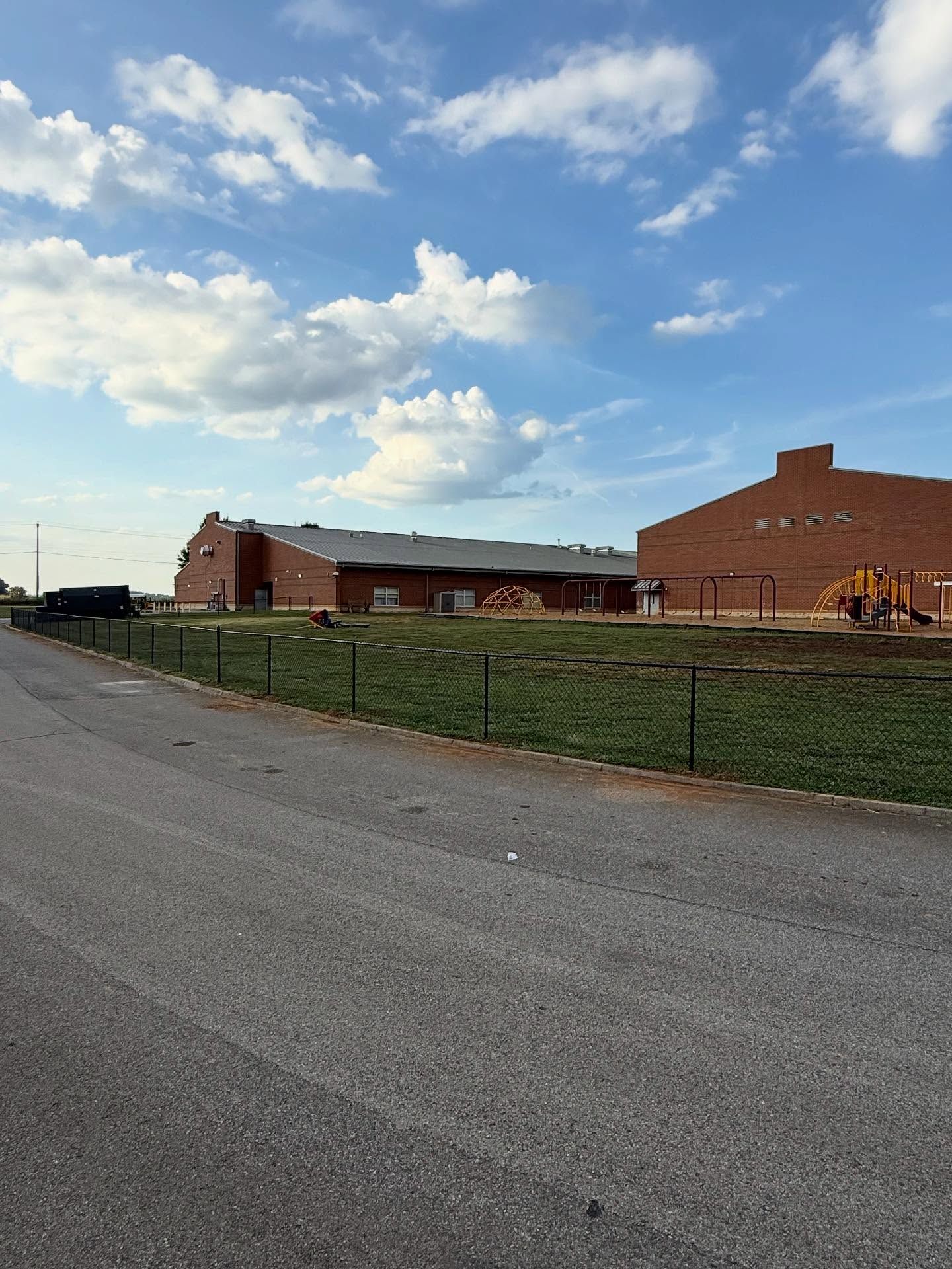 Brick school building beside a road under a partly cloudy blue sky, with grass and a playground nearby