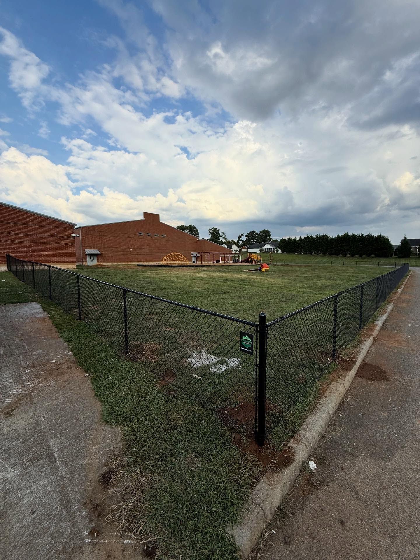 Fenced grassy lot beside brick buildings under a cloudy sky
