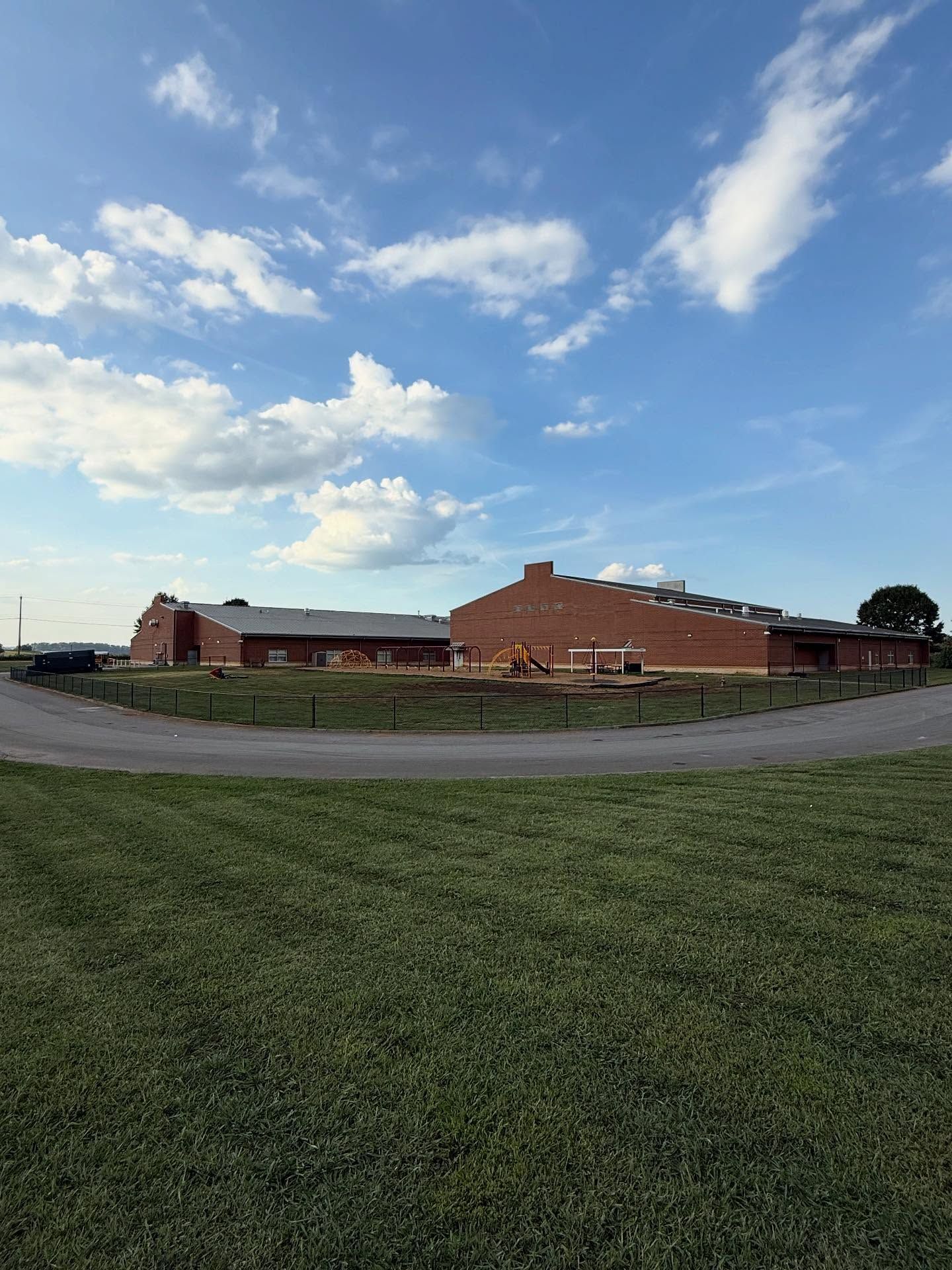 Red brick buildings beside a grassy field under a blue sky with scattered clouds.