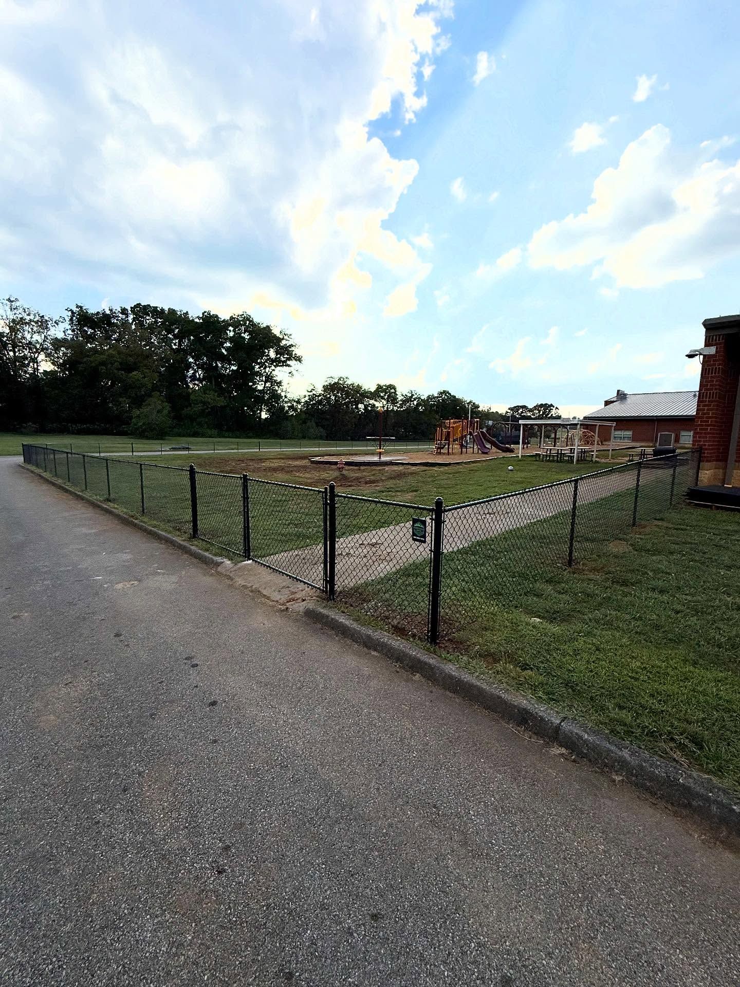 Gravel path beside a fenced grassy enclosure at dusk, with trees and a brick building in the background