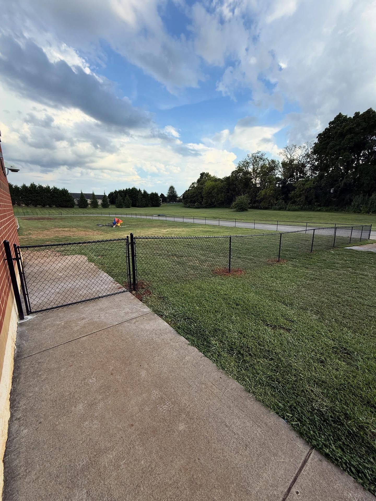 Concrete walkway beside a grassy yard and road under a cloudy blue sky, with a house wall on the left