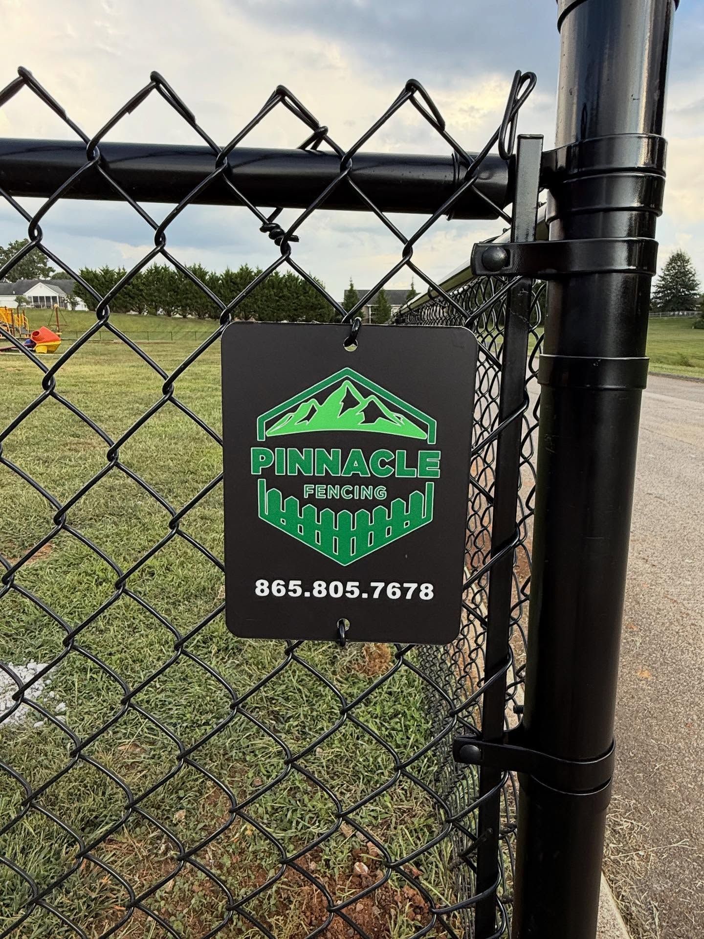 Black chain-link fence sign with green logo and phone number on a sports field fence.