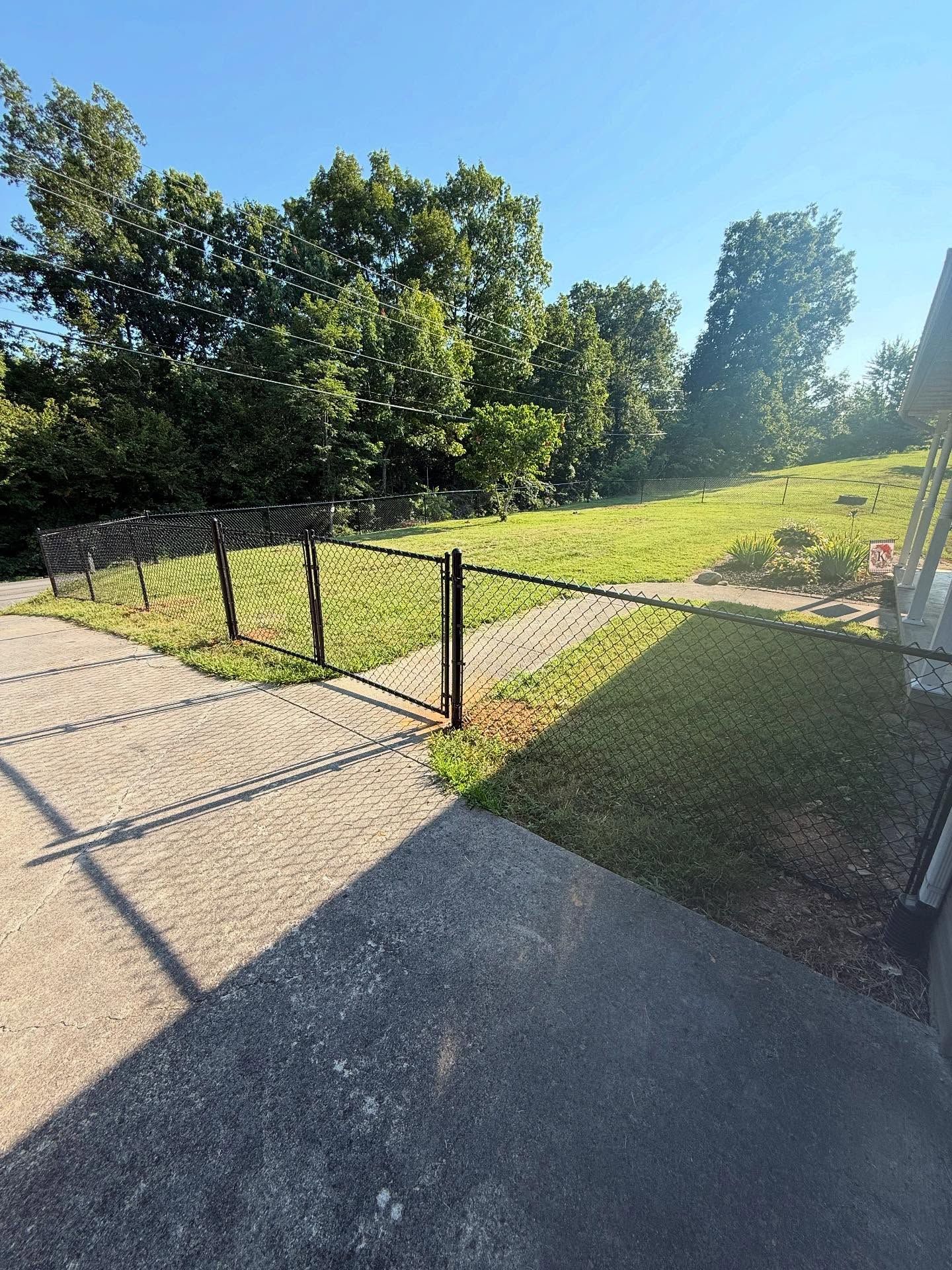 Sunlit paved path beside a grassy slope and black fence, with trees and bright sky in the background