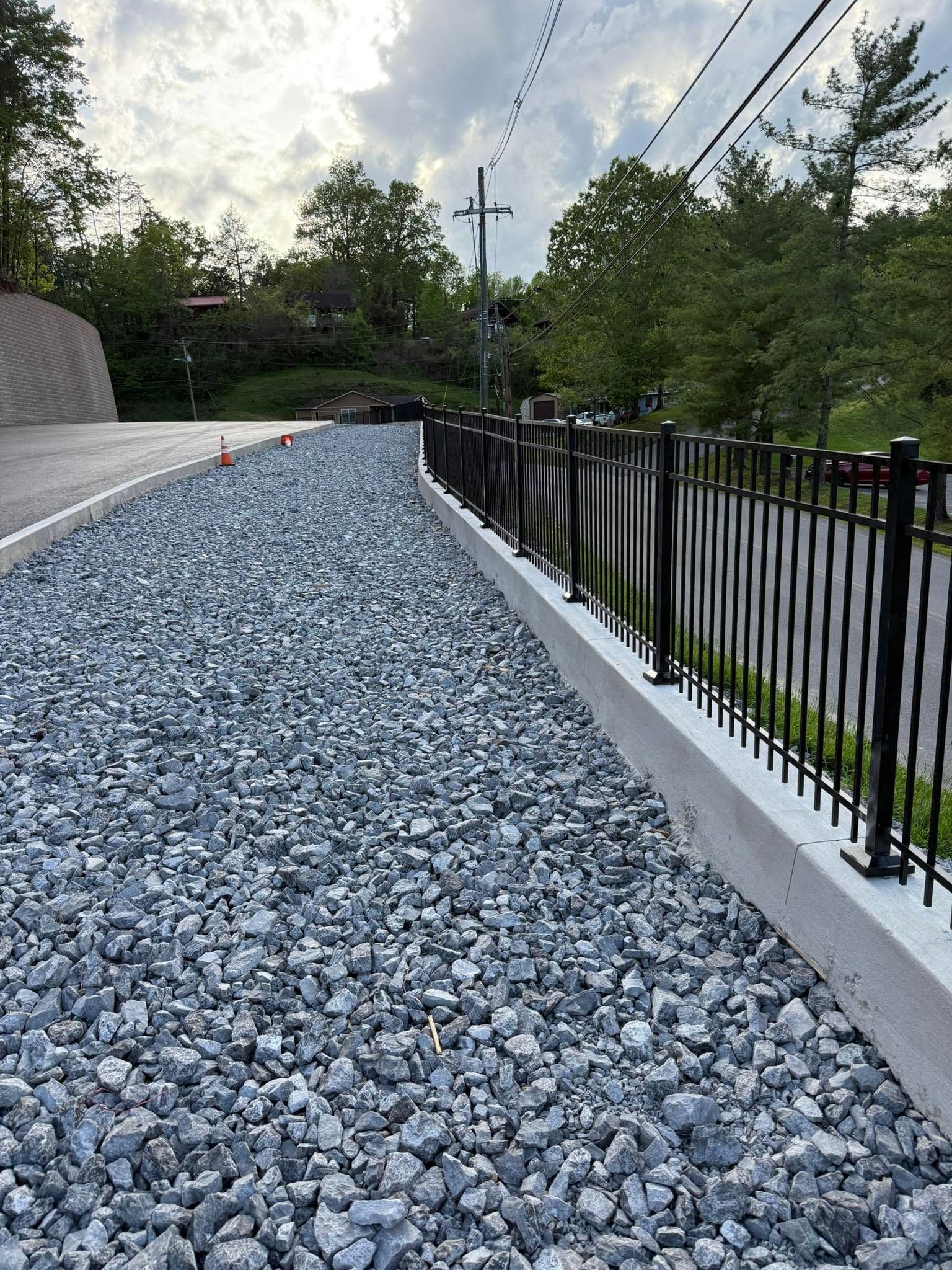 Gravel-covered sloped walkway beside a black fence and trees under a cloudy sky