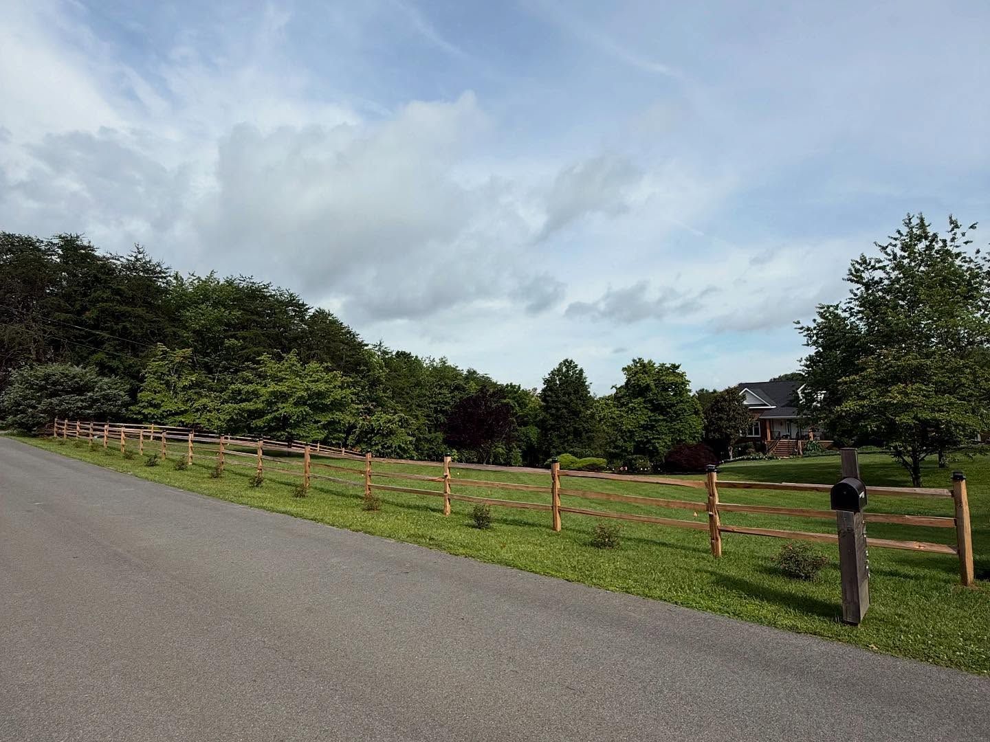 Curving paved road beside a wooden fence and grassy field under a cloudy sky