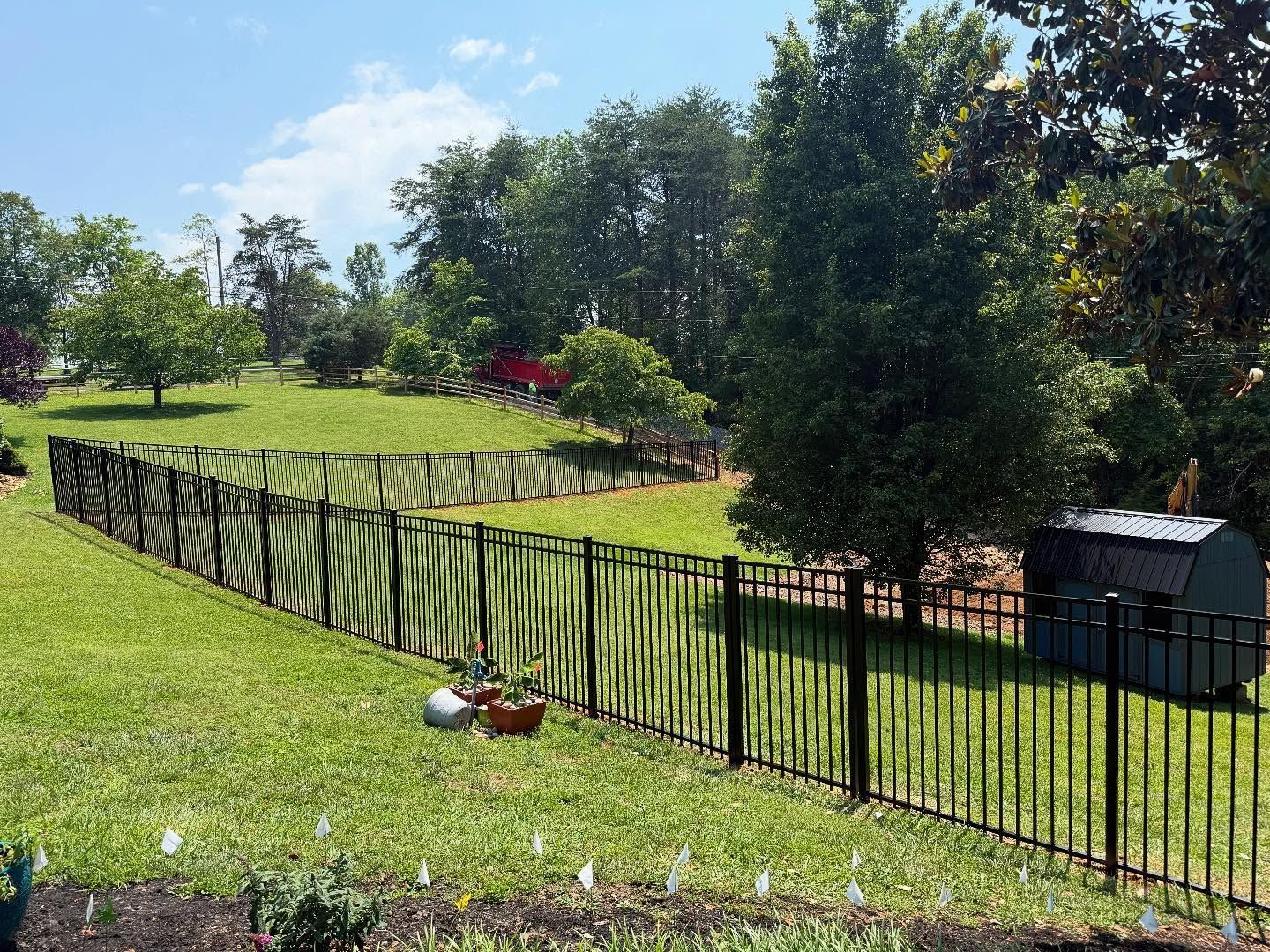 Black metal fence around a grassy yard with trees and a small shed on a sunny day