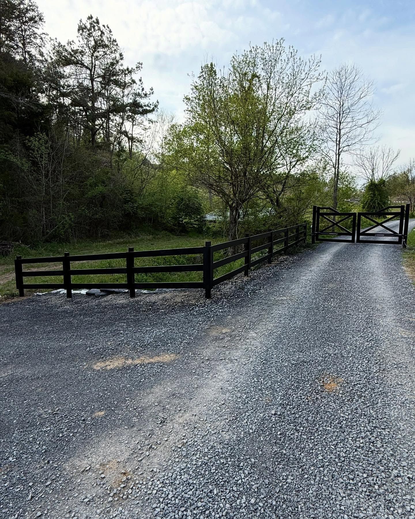 Gravel driveway beside black wooden fence and trees leading to a closed gate