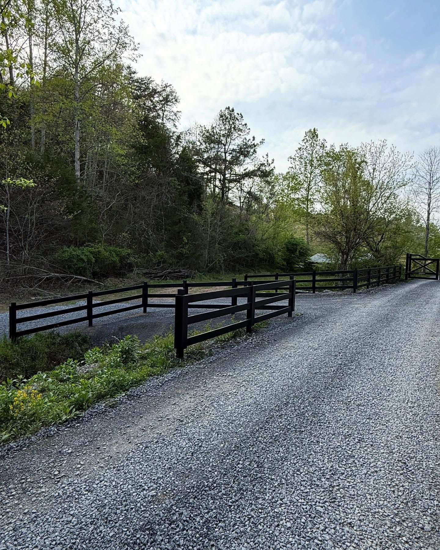 Gravel road beside a black wooden fence and trees under a bright sky