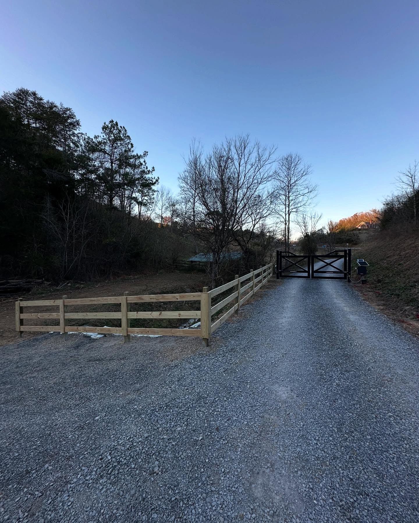 Gravel driveway leading to a wooden gate at dusk, bordered by a fenced grassy area and trees.