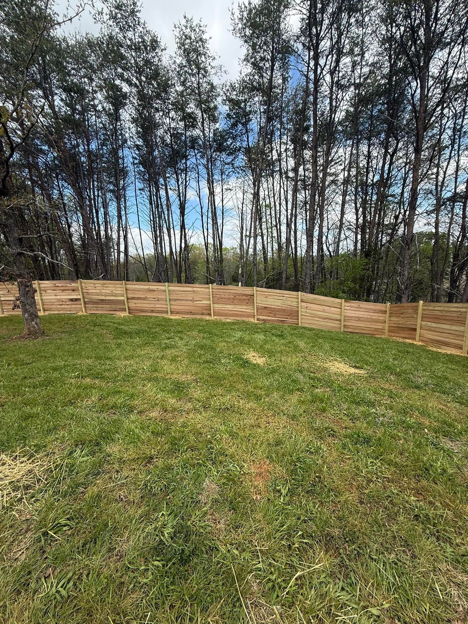 Wooden fence along a grassy hill with tall trees and blue sky in the background
