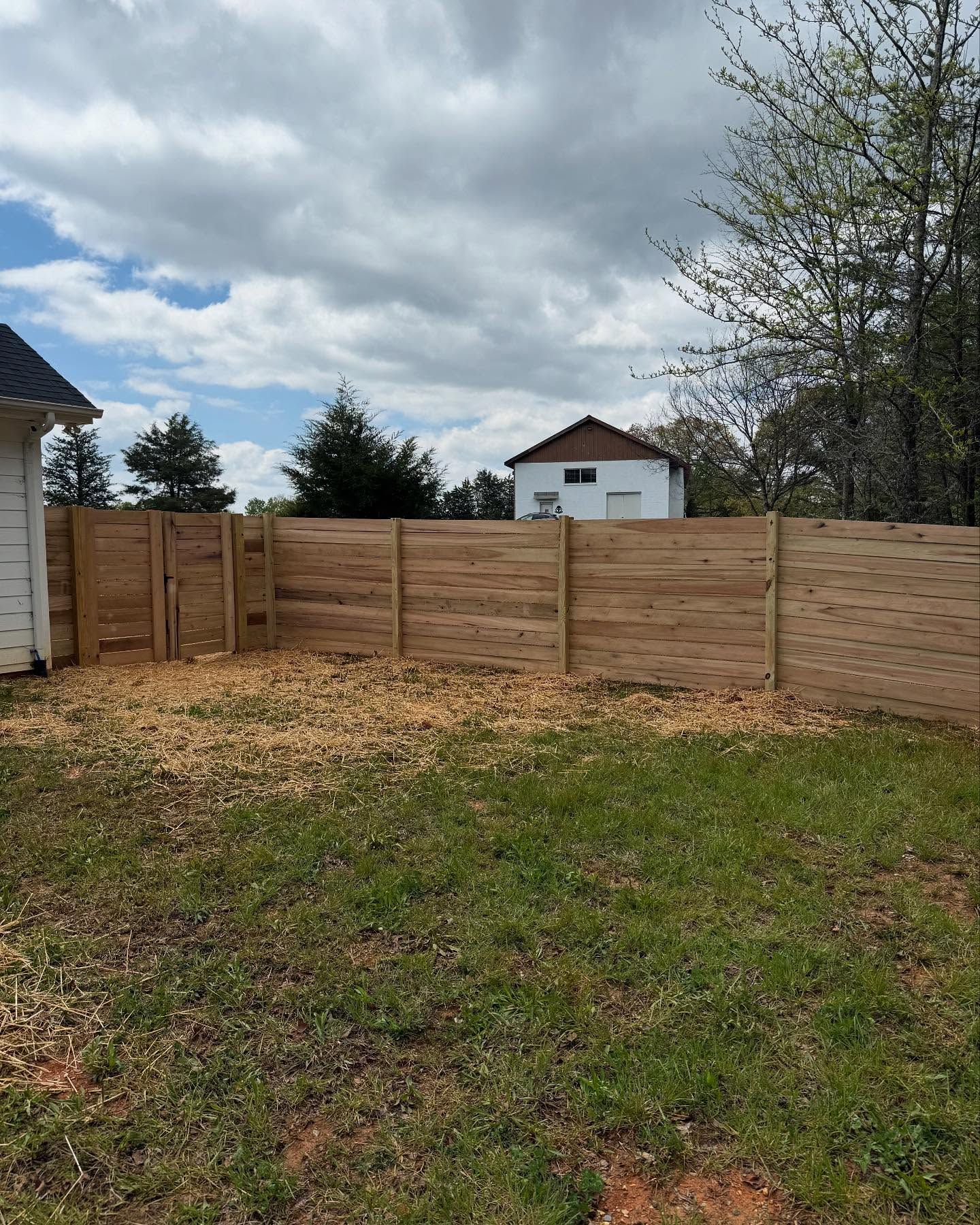 Wooden privacy fence in a grassy yard under a cloudy sky, with houses and trees in the background.