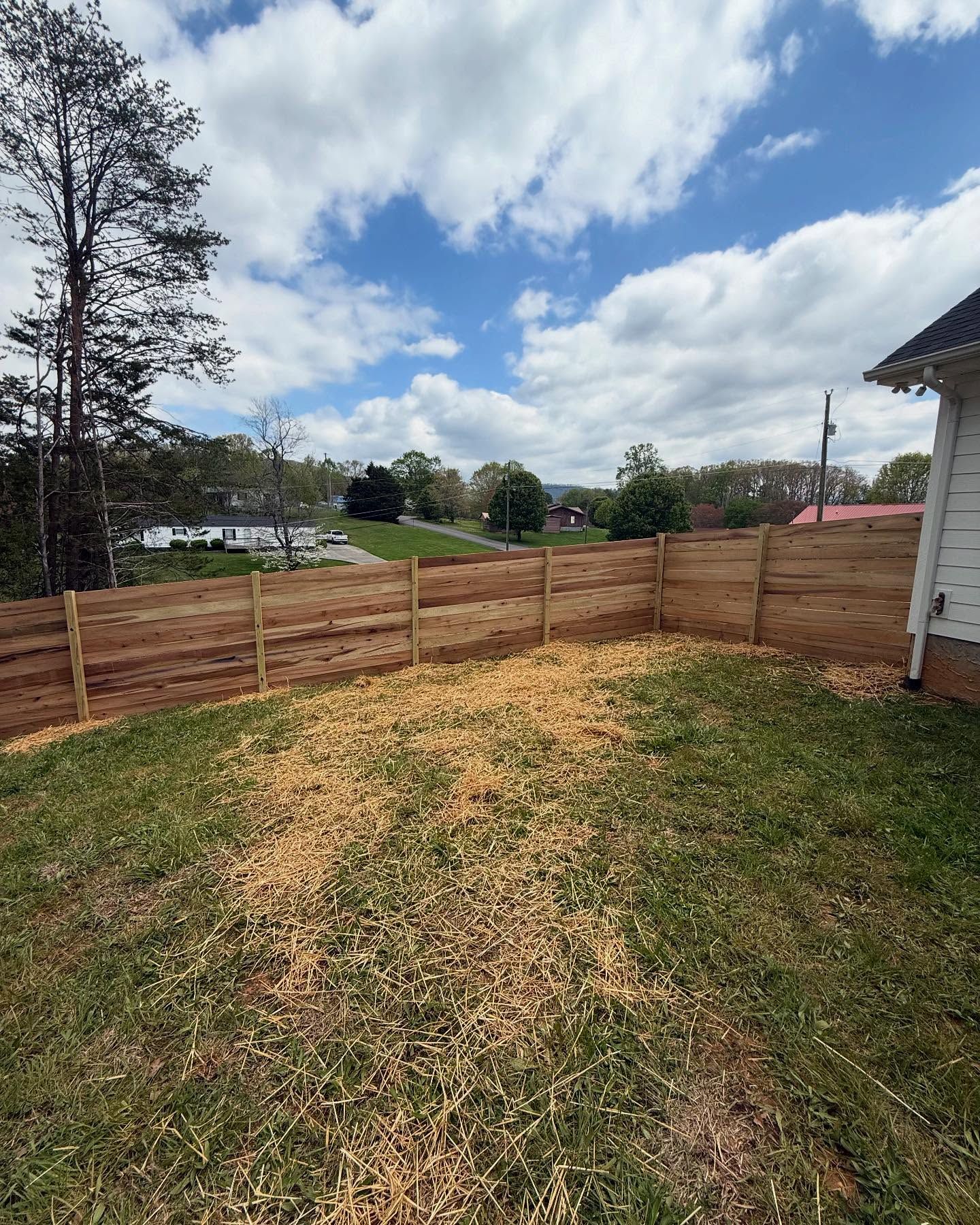 Backyard with a wooden fence, patchy grass, and cloudy sky beside a house